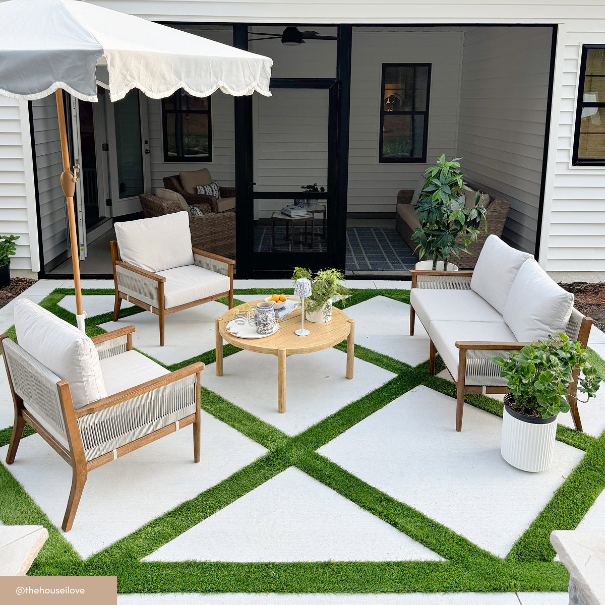 Modern outdoor patio with white cushioned wooden chairs and a sofa around a round wooden table, set on a geometric white and green grass-patterned floor, next to a house with large glass doors.