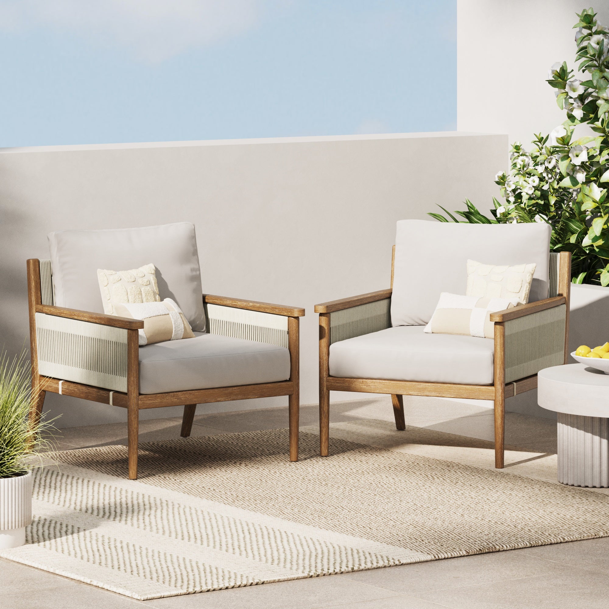 Two modern wooden armchairs with light cushions and decorative pillows sit on a woven rug on a patio, surrounded by green plants and a white table under a blue sky.