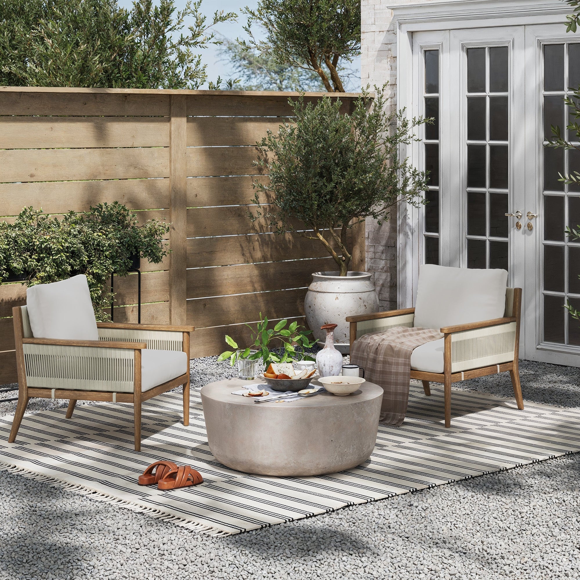 Two Nathan James Outdoor Wood & Rope Patio Arm Chairs in light gray with washable white cushions face a round stone table with drinks and snacks on a striped rug in a sunny gravel patio with plants, wooden fence, and glass doors behind.