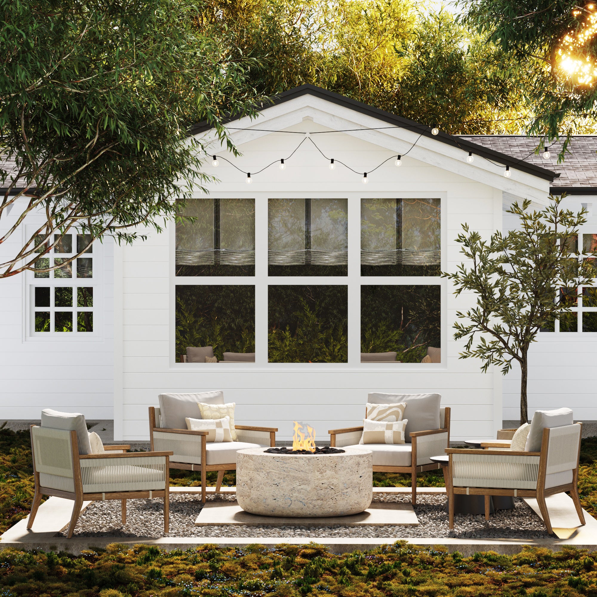 A cozy outdoor patio features cushioned chairs arranged around a stone fire pit, set on a gravel area in front of a white house with large windows and string lights overhead. Trees and greenery surround the space.
