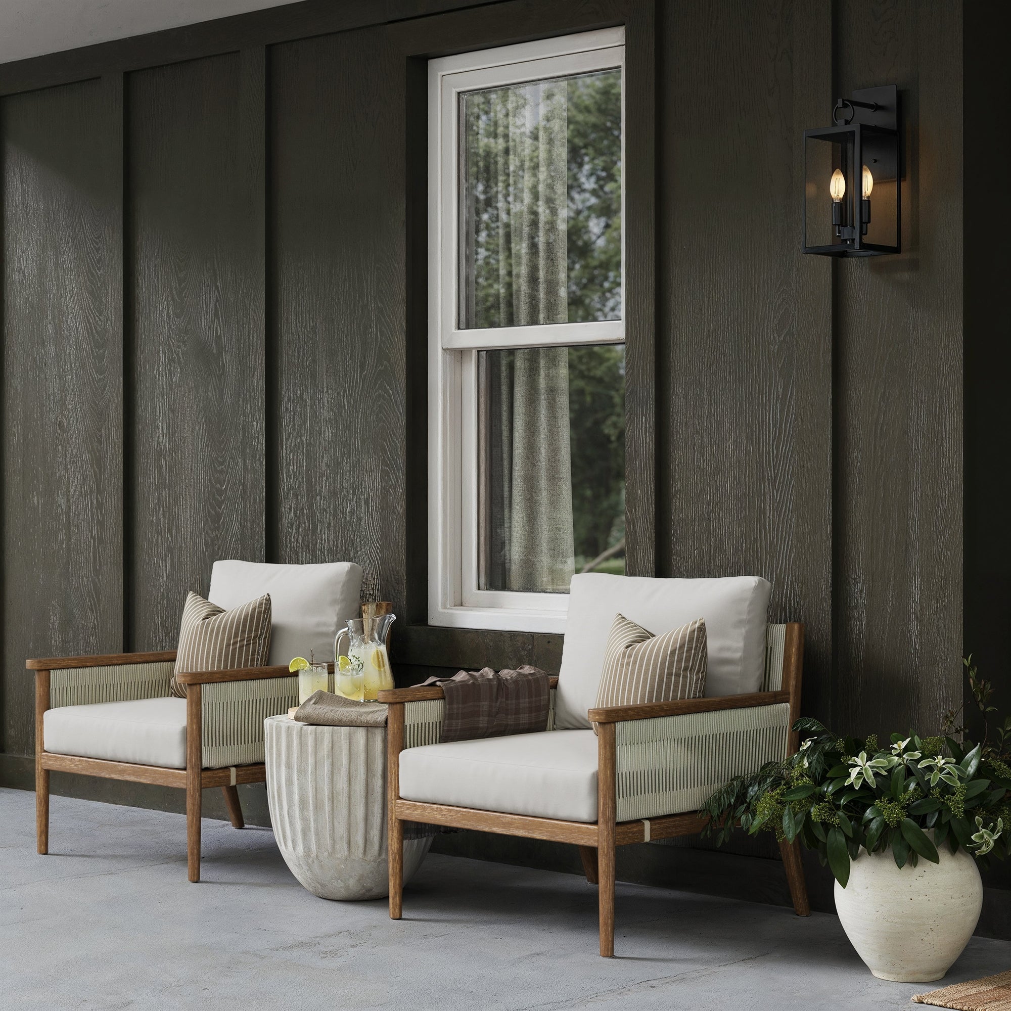 Two wooden chairs with white cushions sit on a covered patio next to a round side table with lemonade. A potted plant is on the ground nearby, and a black wall lantern hangs by a window in a dark wood-paneled wall.