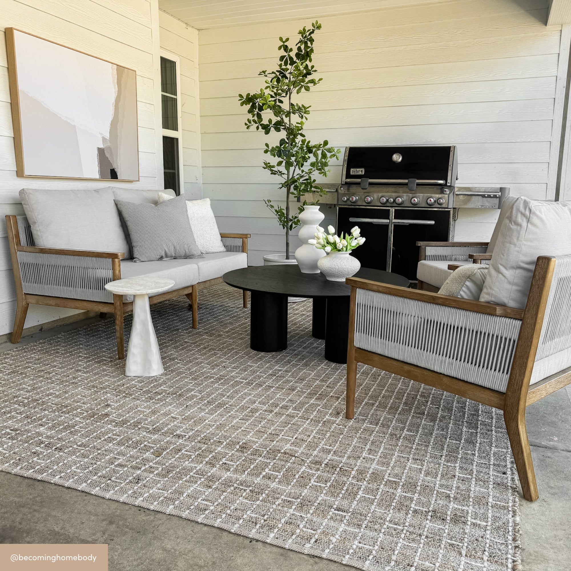 Modern outdoor patio with Nathan James Outdoor Wood & Rope Patio Arm Chairs in light gray (set of 4), a sofa with removable washable covers, round black coffee tables, grill, potted plants, wall art, and a textured rug under covered white paneled walls.