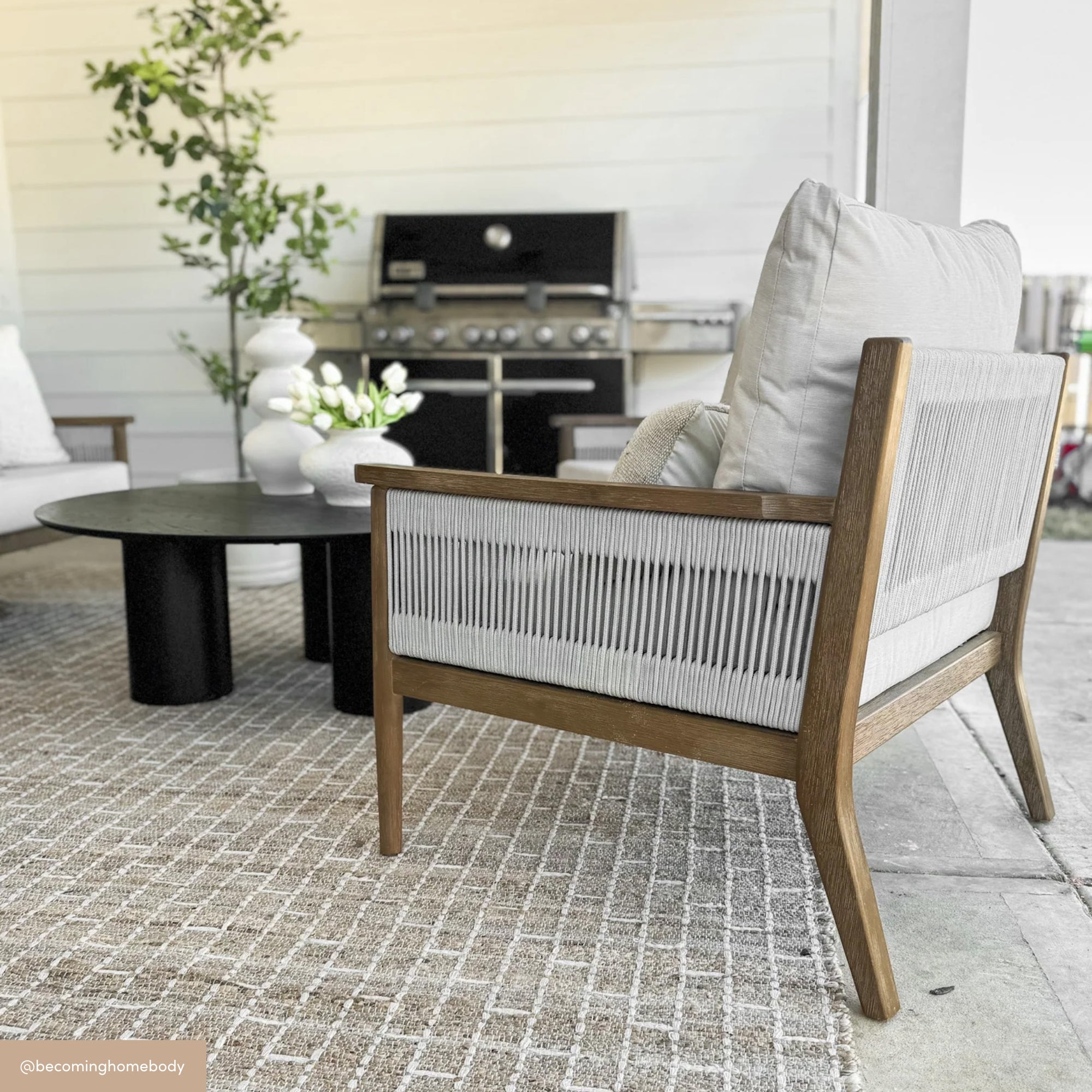 Modern outdoor patio with a woven armchair, cushioned seat, round black coffee table decorated with white vases and flowers, and a grill in the background on a patterned rug.