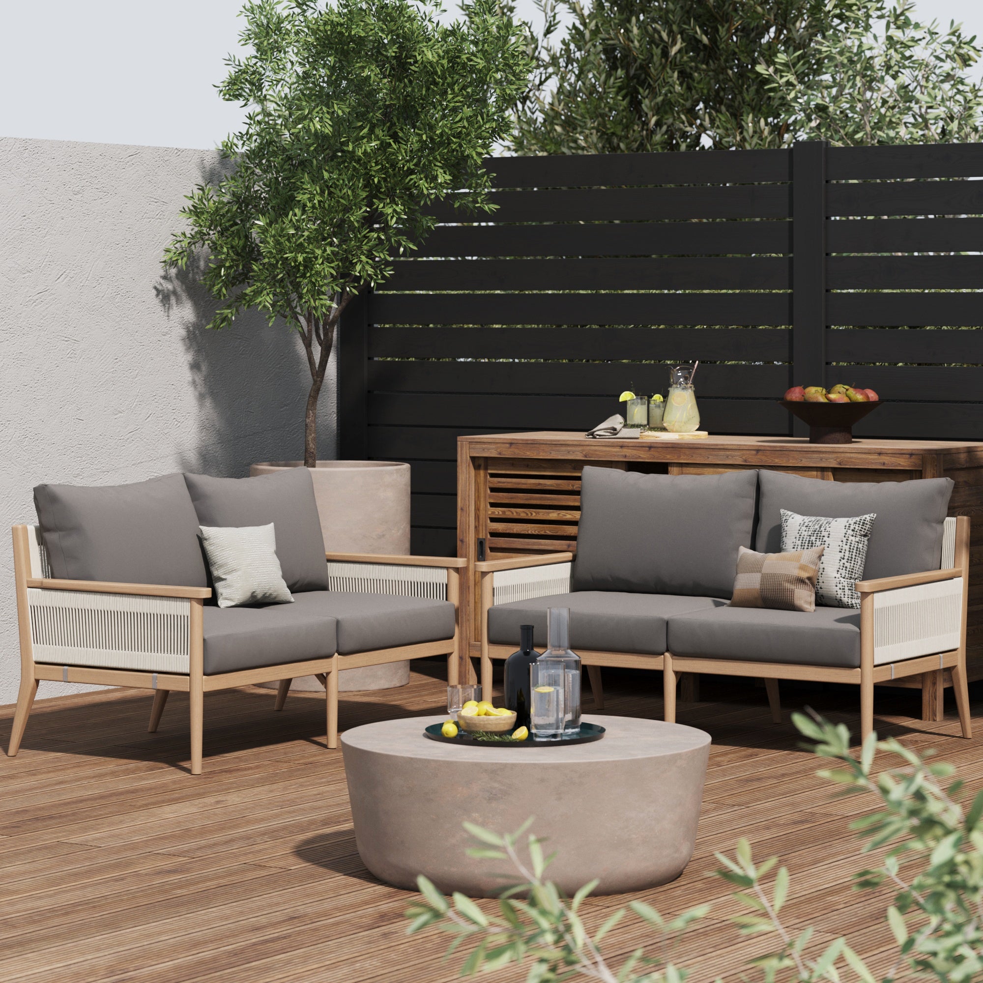 Modern patio with Nathan James Outdoor Wood & Rope Patio Loveseats in medium gray, gray weather-resistant cushions, a round coffee table with drinks and lemons, lush green plants, and a sleek black privacy fence.