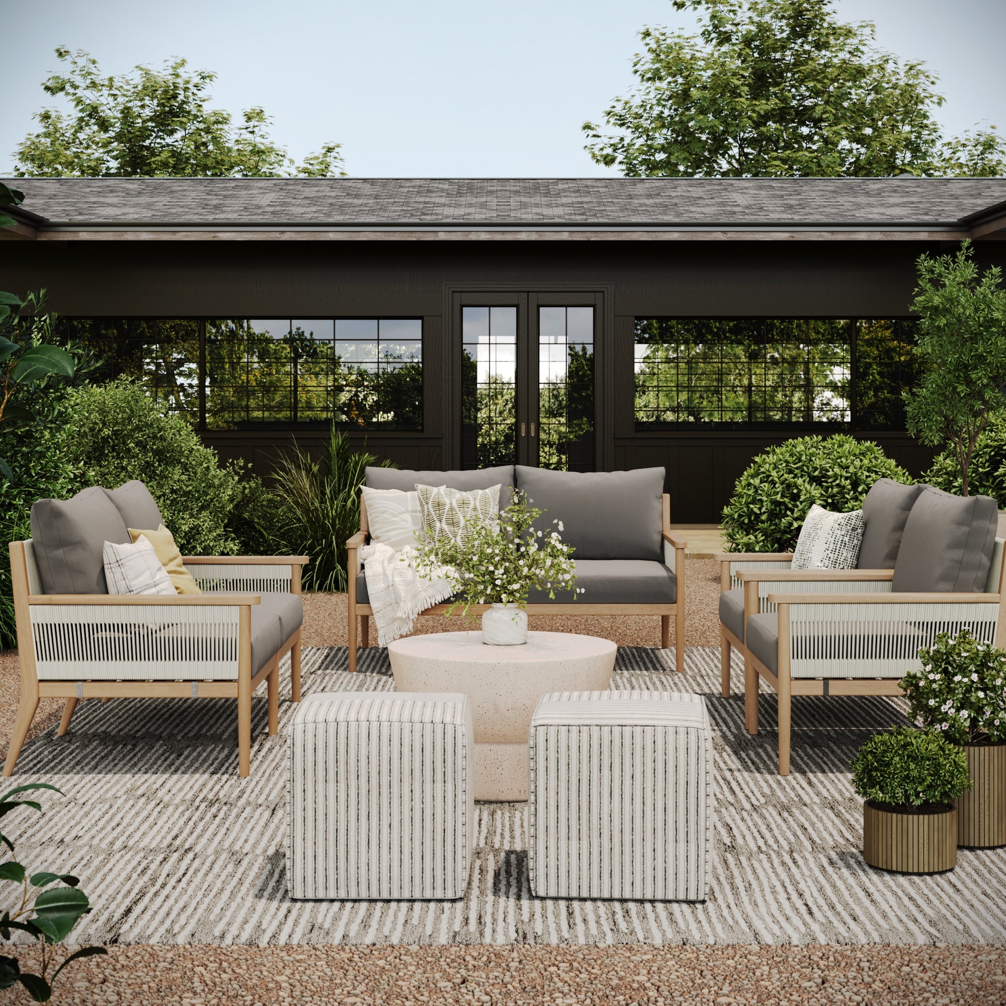 A stylish modern patio featuring the Nathan James Outdoor 4 Piece Wood Patio Set Loveseats and Chairs with gray washable cushions, striped ottomans, a round coffee table with flowers, green plants, and a striped rug by a dark building.