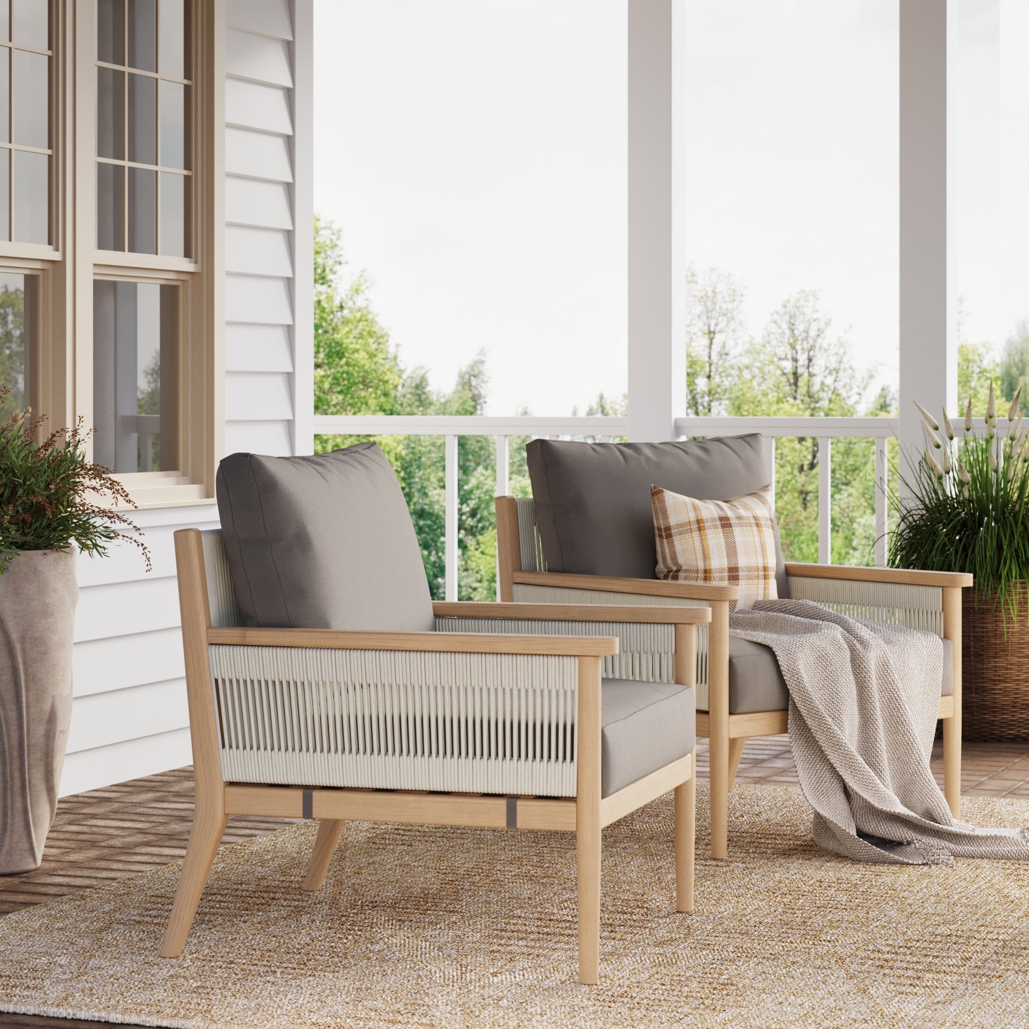 Two Nathan James Outdoor Wood & Rope Patio Arm Chairs in medium gray sit on a porch—one with a plaid pillow, the other with a beige throw. Gray removable cushion covers, potted plants, and a railing overlook green trees in the background.