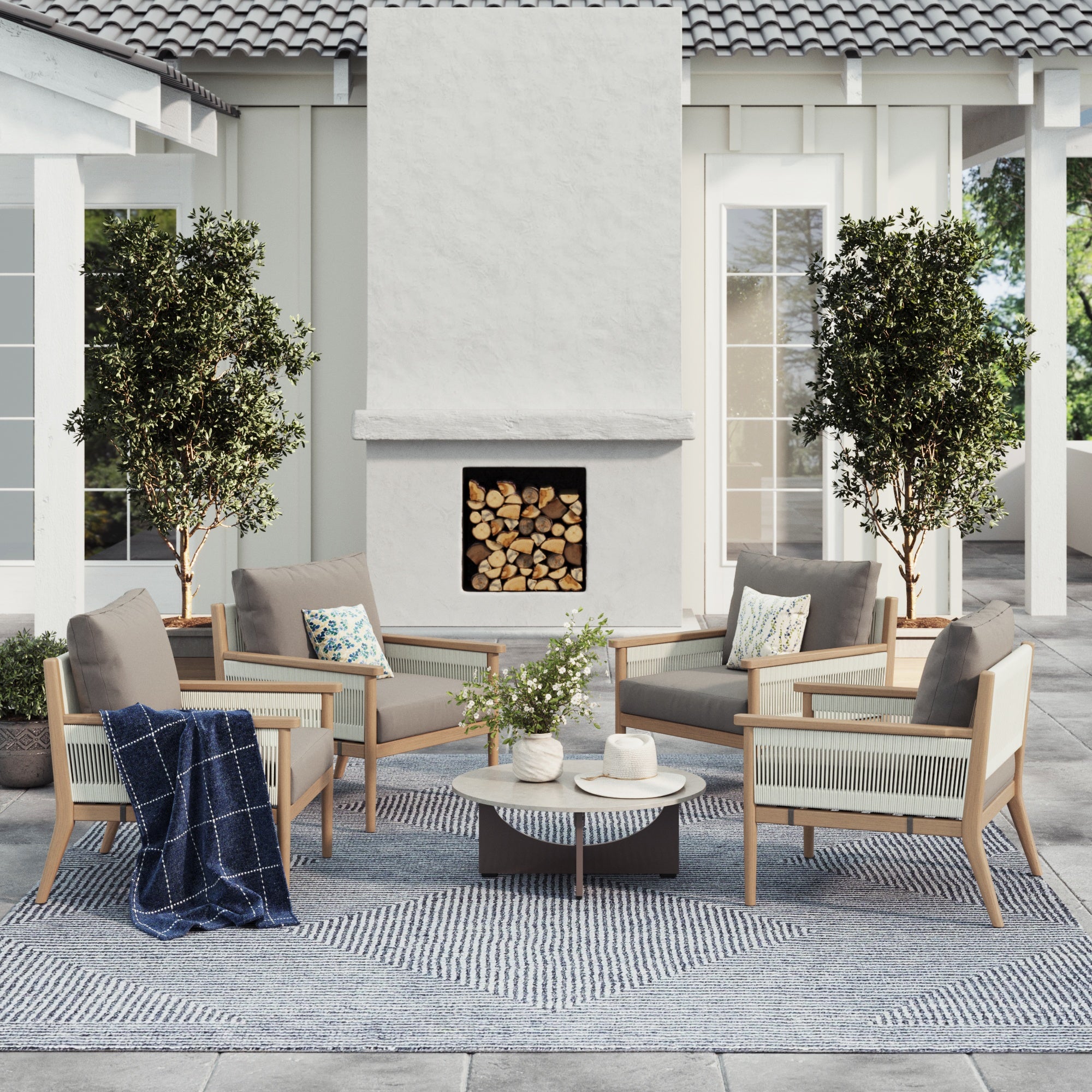 A modern patio features four Nathan James Outdoor Wood & Rope Patio Arm Chairs in medium gray around a round coffee table, with a fireplace, two potted trees, a geometric rug, and cozy touches like a blanket and pillows.