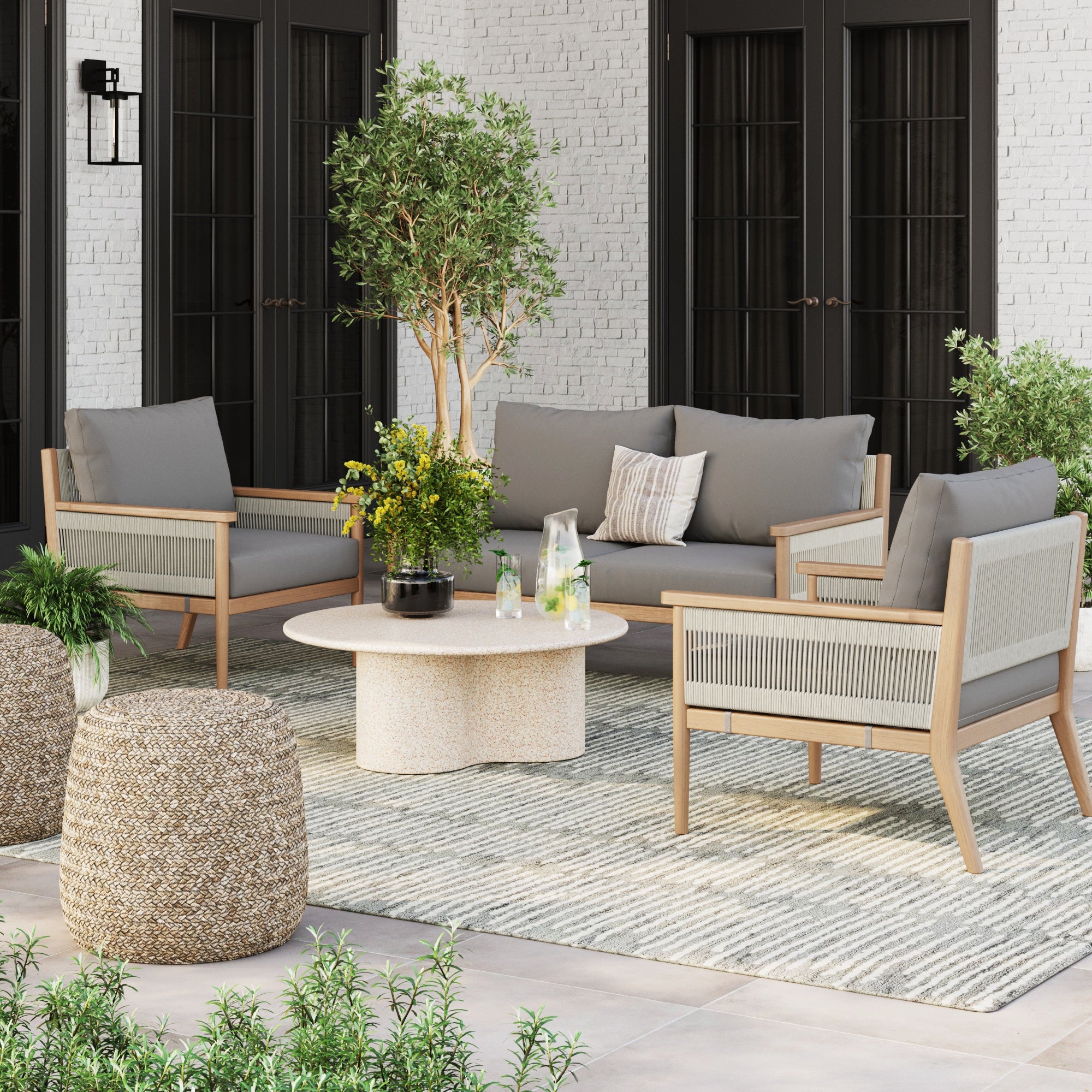 A stylish patio features a gray sofa, two Nathan James Outdoor Wood & Rope Patio Arm Chairs in medium gray with washable cushion covers, a round table with drinks and flowers, a woven pouf, potted plants, and a striped rug on tiled floor by white brick walls.