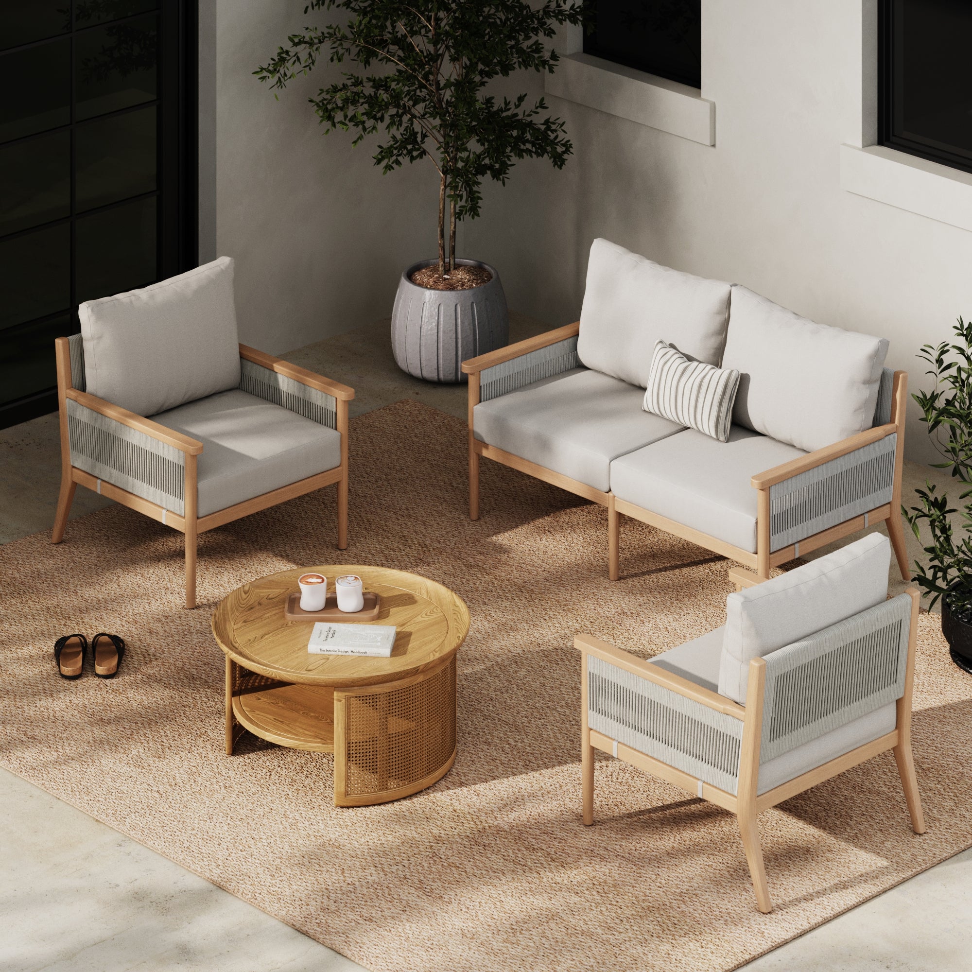 A modern outdoor seating area includes the Nathan James Outdoor 3 Piece Wood Patio Set Loveseat and Chairs in white, a round wooden coffee table, potted plants, a woven rug, and sandals on a concrete patio.