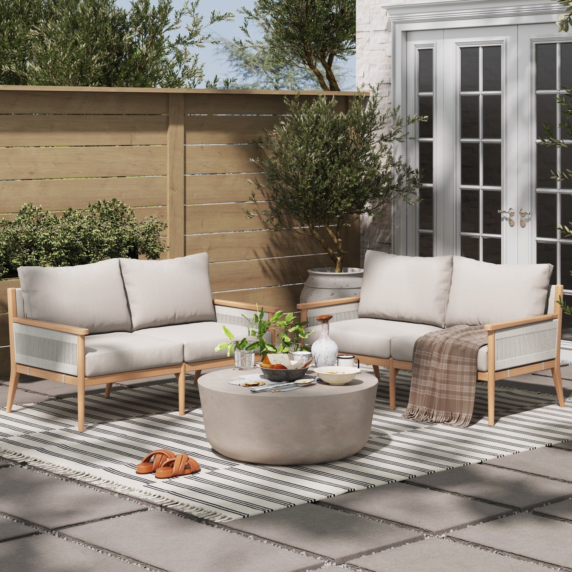 A cozy patio with two Nathan James Outdoor Wood & Rope Patio Loveseats Ash White, weather-resistant cushions, a round coffee table, striped rug, potted plants, throw blanket, tray with bottles and glasses, and brown sandals by French doors.