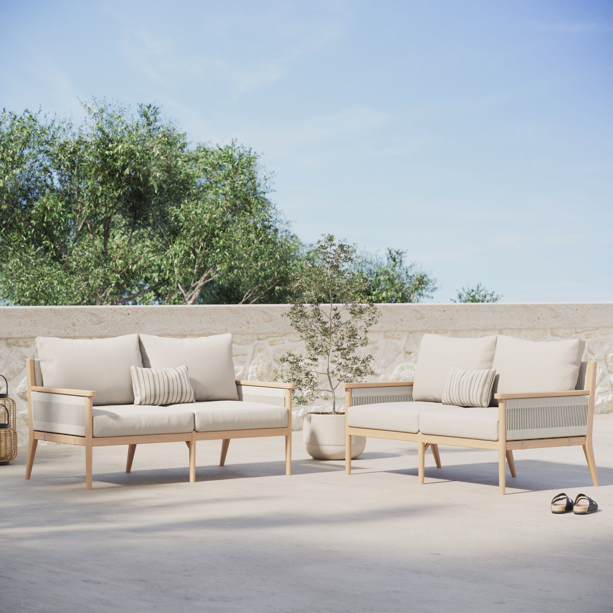 Two Nathan James Outdoor Wood & Rope Patio Loveseats in Ash White, featuring weather-resistant cushions and striped pillows, are arranged on a patio with a potted plant between them, all under green trees and blue sky.