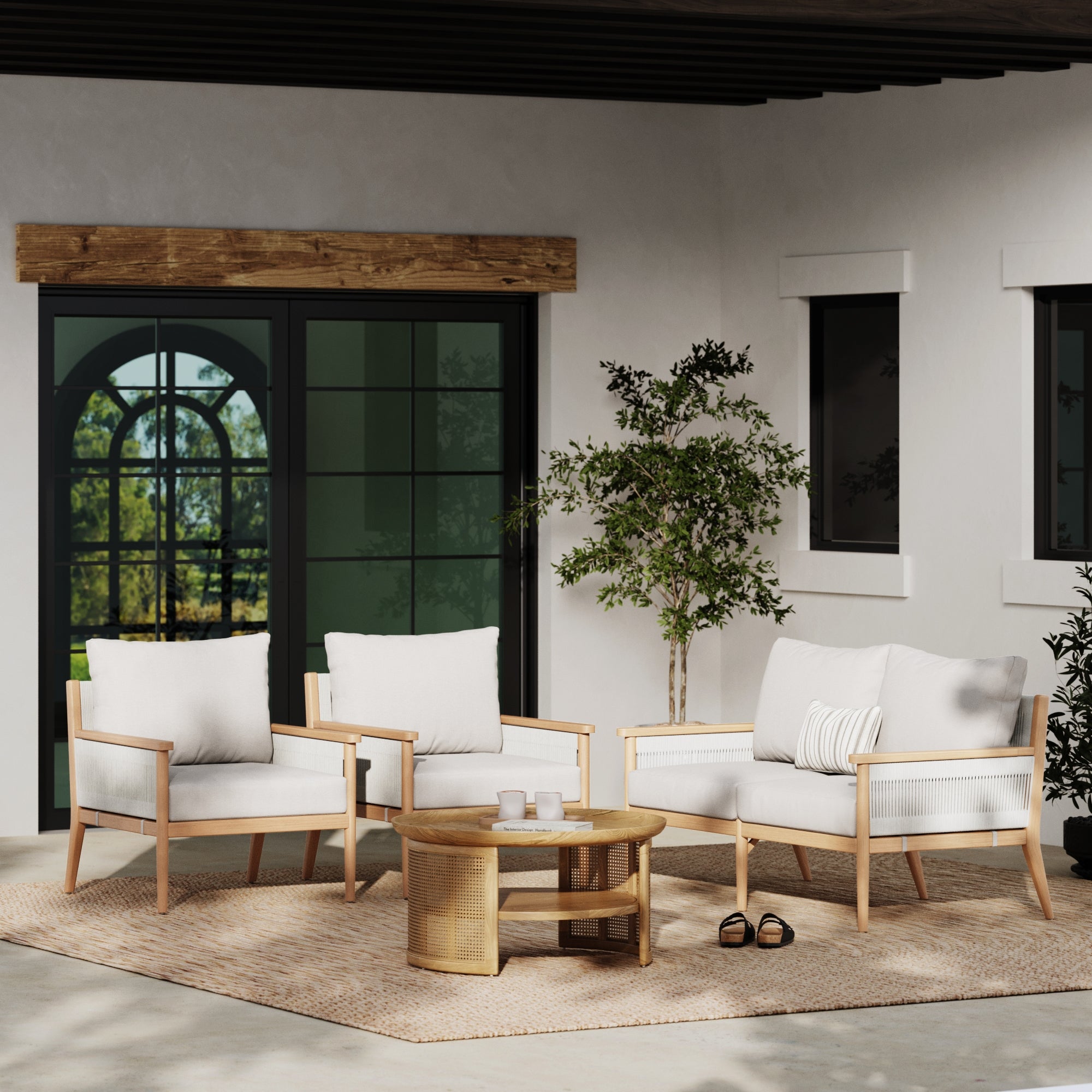 A modern patio features two armchairs and a Nathan James Outdoor Wood & Rope Patio Loveseat in Ash White with weatherproof frames, white washable cushions, a round rattan coffee table, potted tree, woven rug, and black-framed windows and doors.