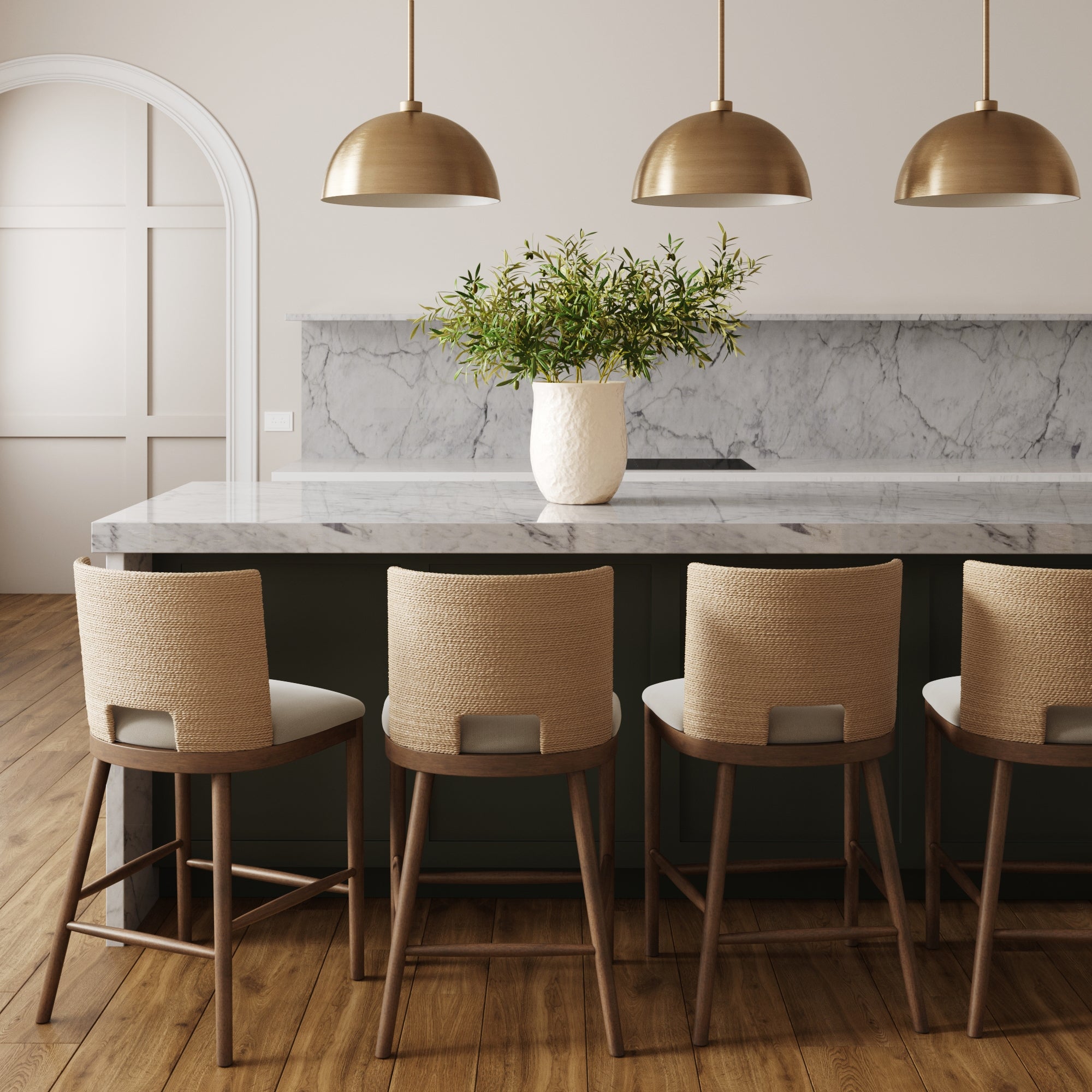 A modern kitchen with a marble island, four Nathan James Seagrass Rope Back Counter Bar Stools in medium brown, three brass pendant lights, and a white vase with greenery. Wooden floors and light-hued walls complete the look.