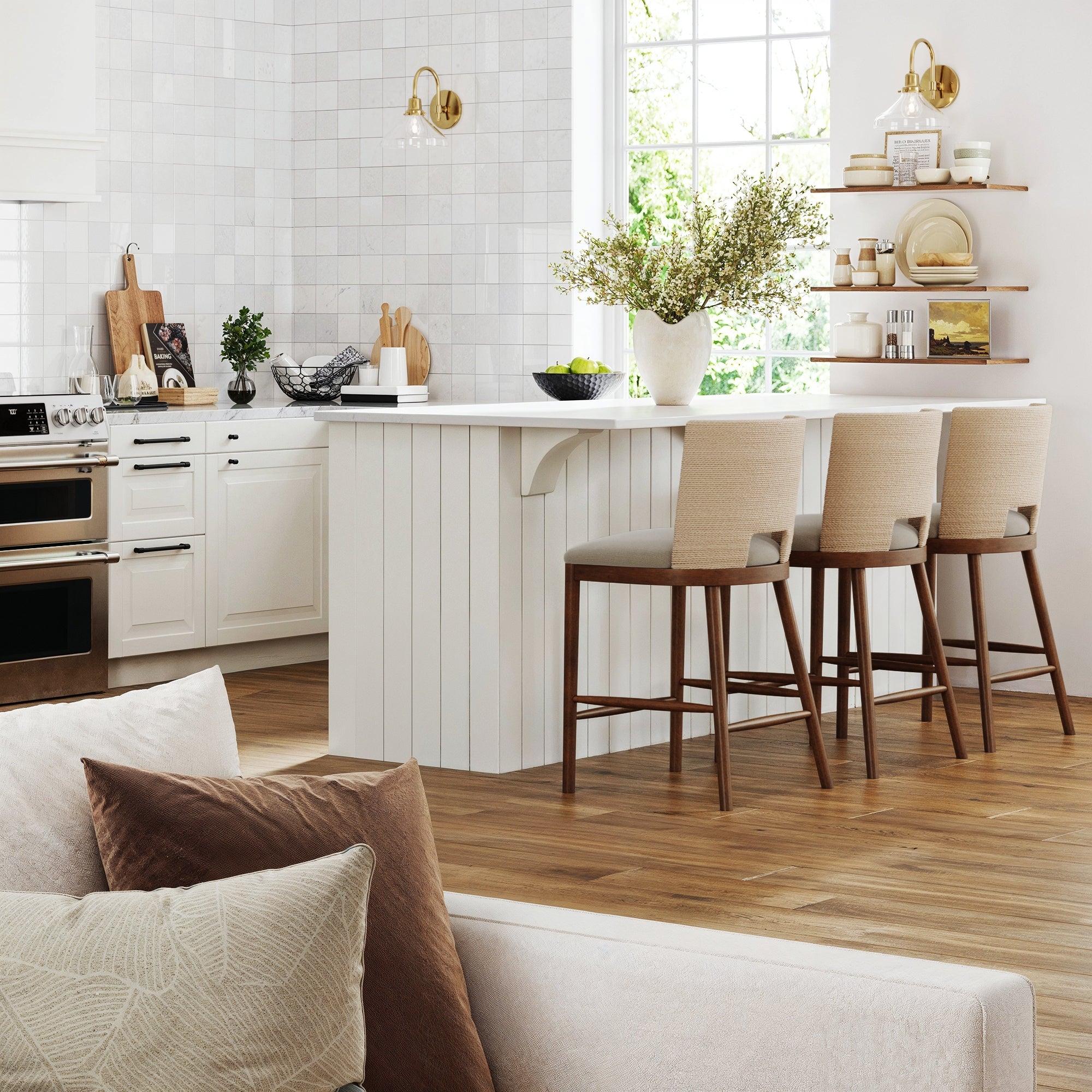Modern kitchen with white cabinets, a paneled white island, three Nathan James Seagrass Rope Back Counter Bar Stools (medium brown), open wood shelves, greenery in a vase, wooden floor, and a sofa with neutral pillows in the foreground.