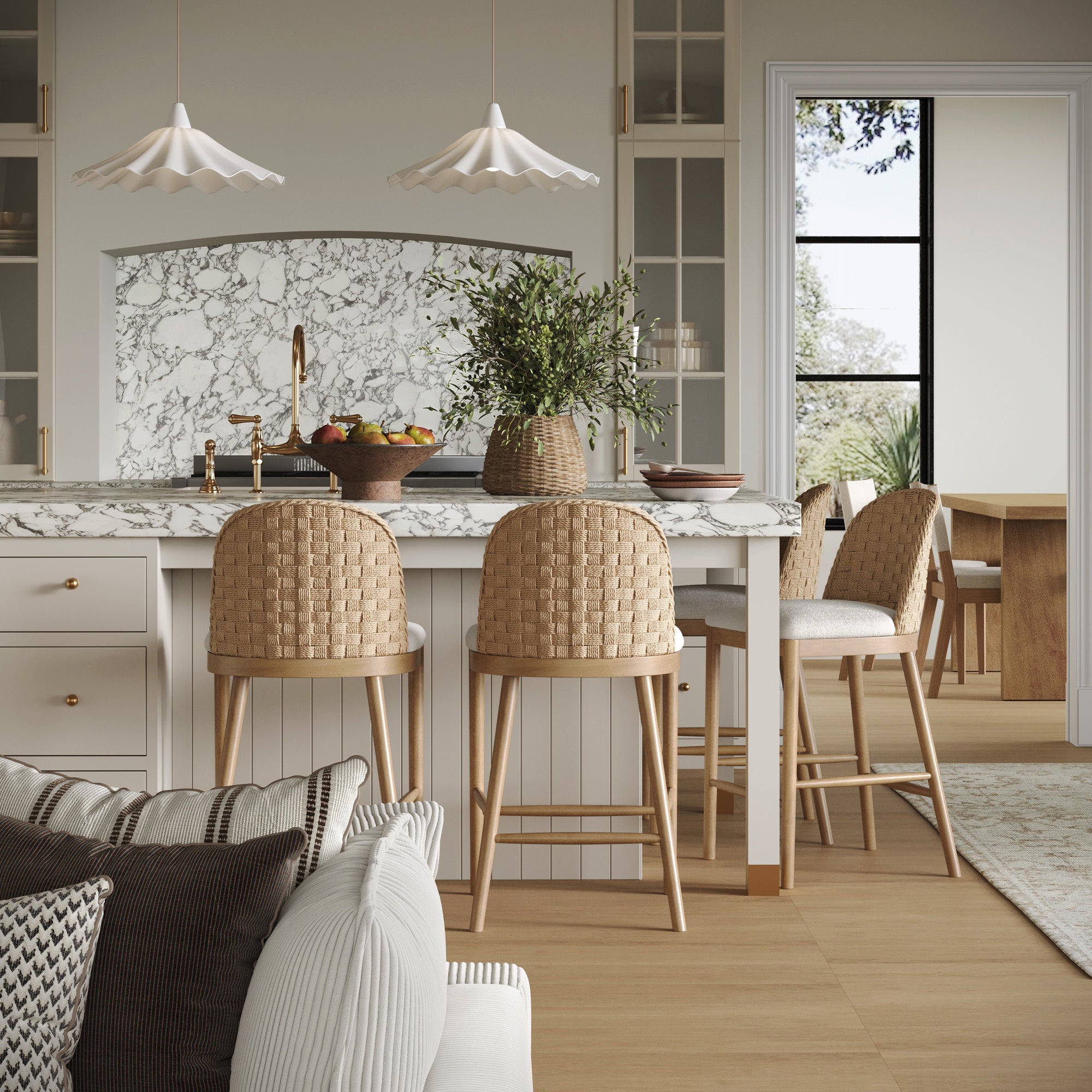 A bright, modern kitchen features a marble island and backsplash, pendant lights, greenery, and Nathan James Woven Seagrass Back & Boucle Counter Bar Stools (Set of 4). Large windows fill the space with light; a cozy living area is visible in front.