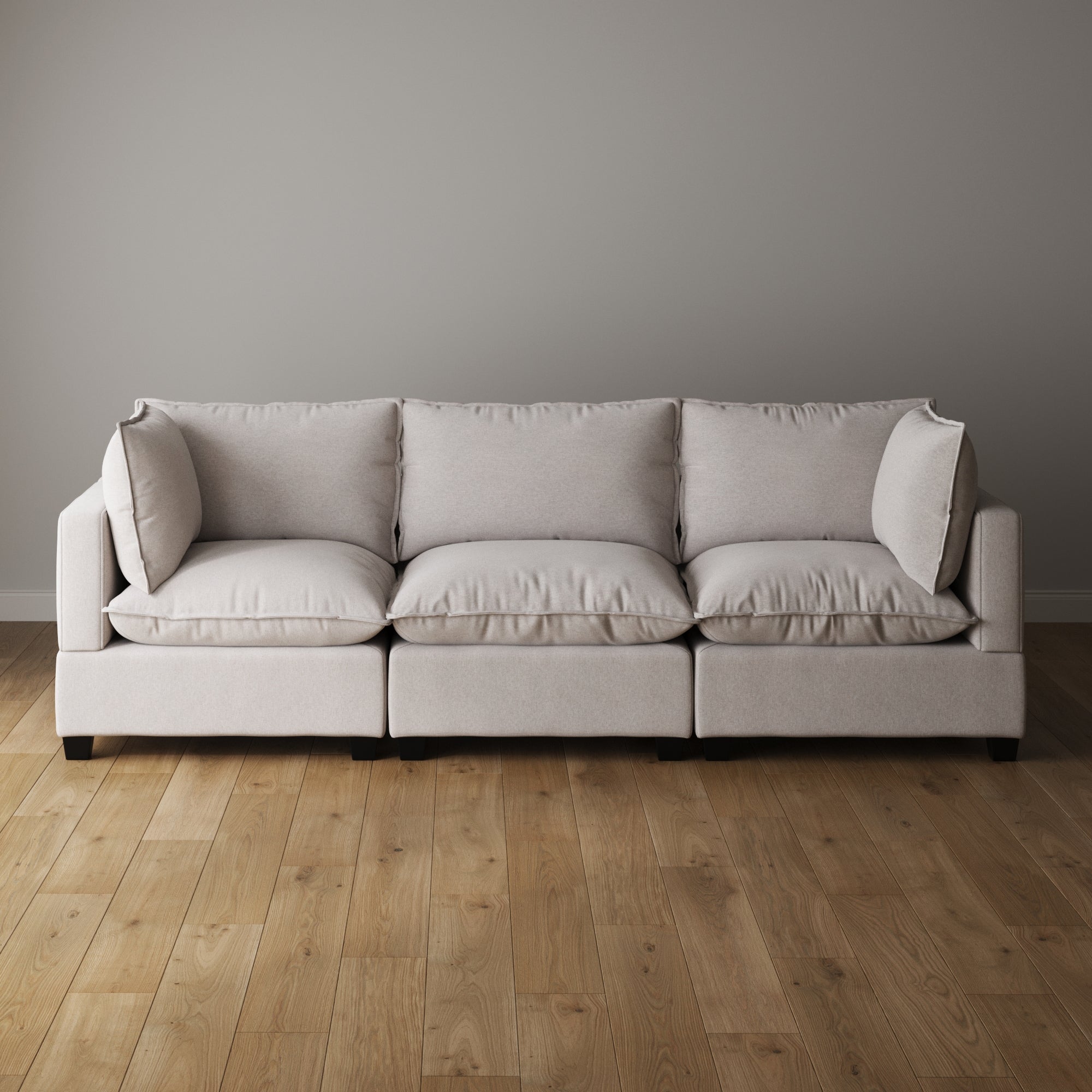 A modern light gray sofa with three seat cushions and three back cushions sits against a plain gray wall on a wooden floor in a minimalist room.