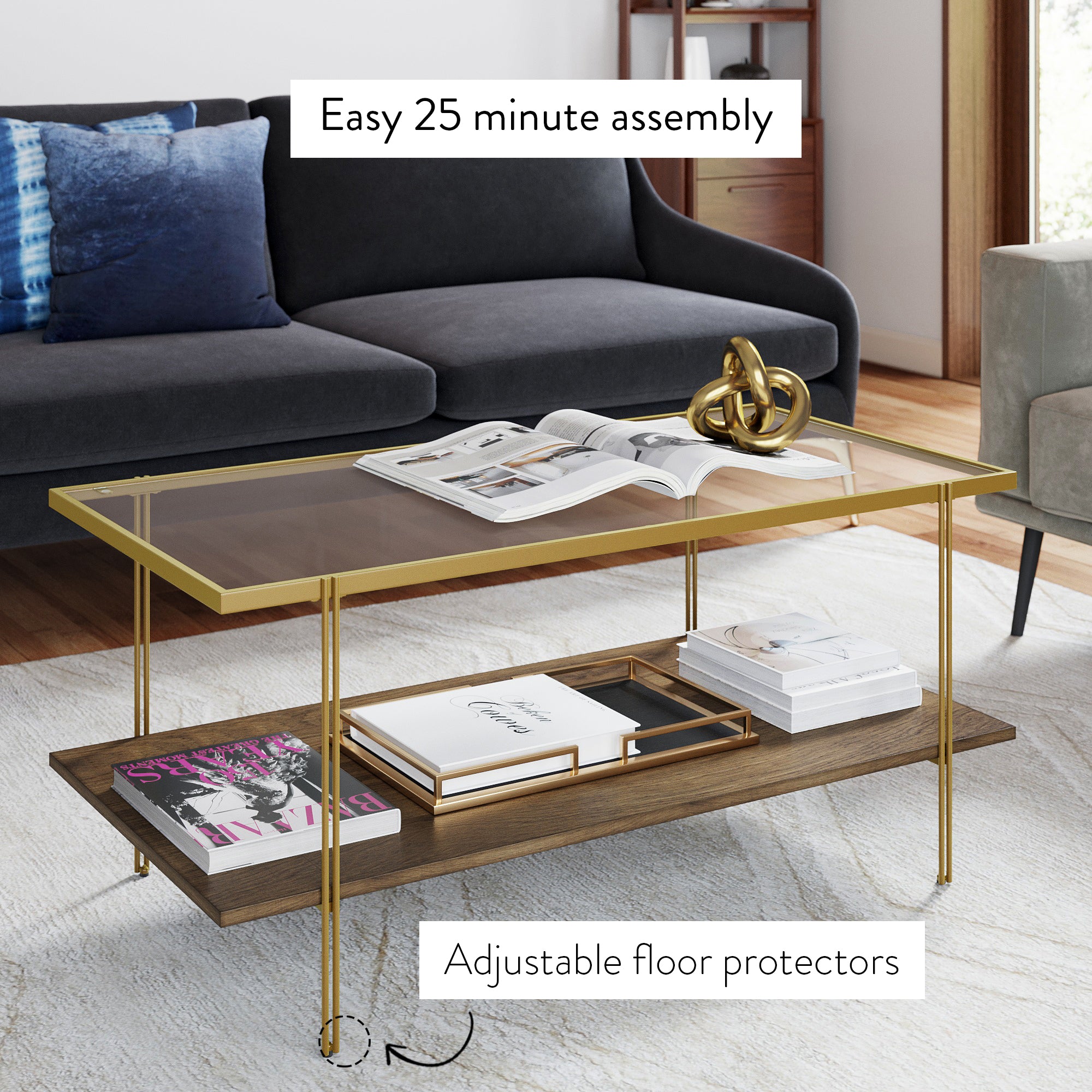 A modern living room with a grey sofa, gold-framed glass coffee table displaying books and decor, and a wooden lower shelf with magazines. Text reads Easy 25 minute assembly and Adjustable floor protectors.
