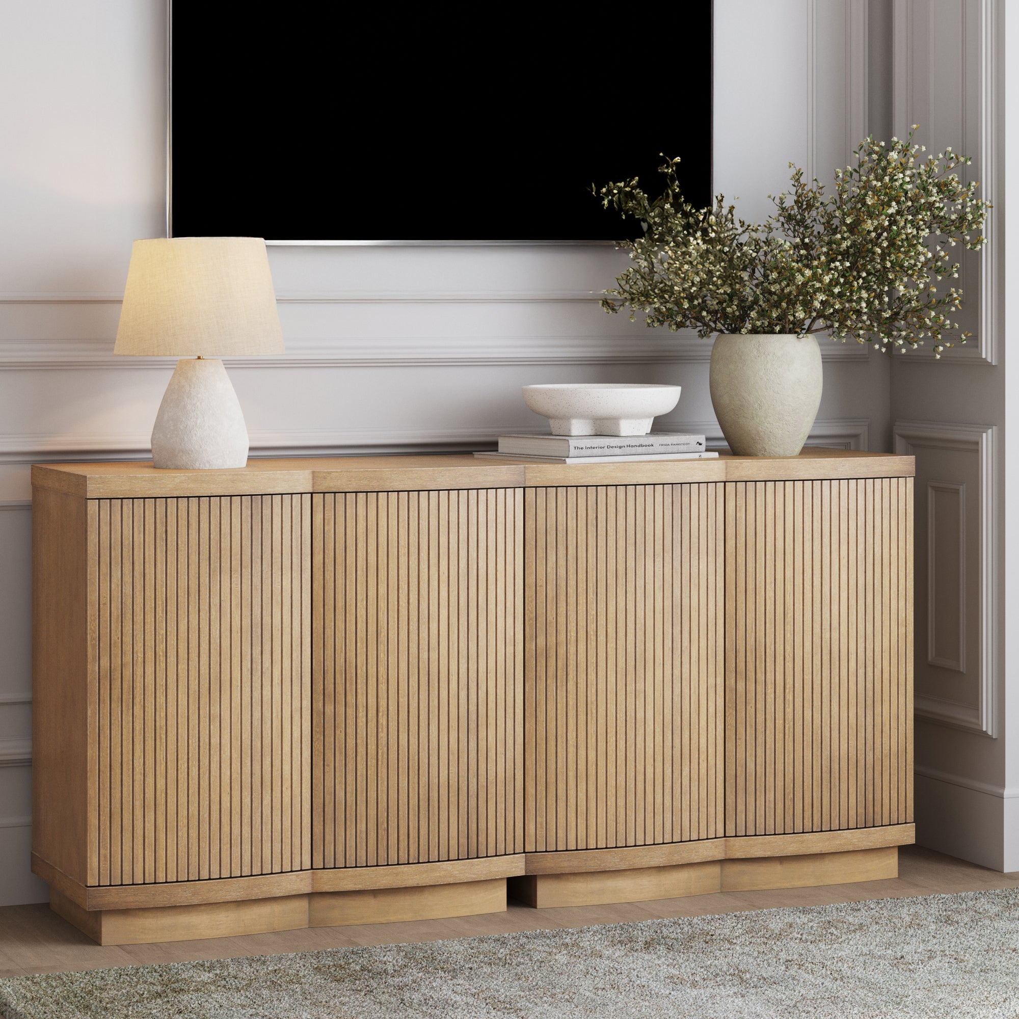 The Nathan James Mid-Century Modern Fluted Console or TV Stand in light brown (set of 2) stands against a white wall, topped with a lamp, stacked books, a white bowl, and greenery in a large vase. Above hangs a black rectangle resembling a TV.