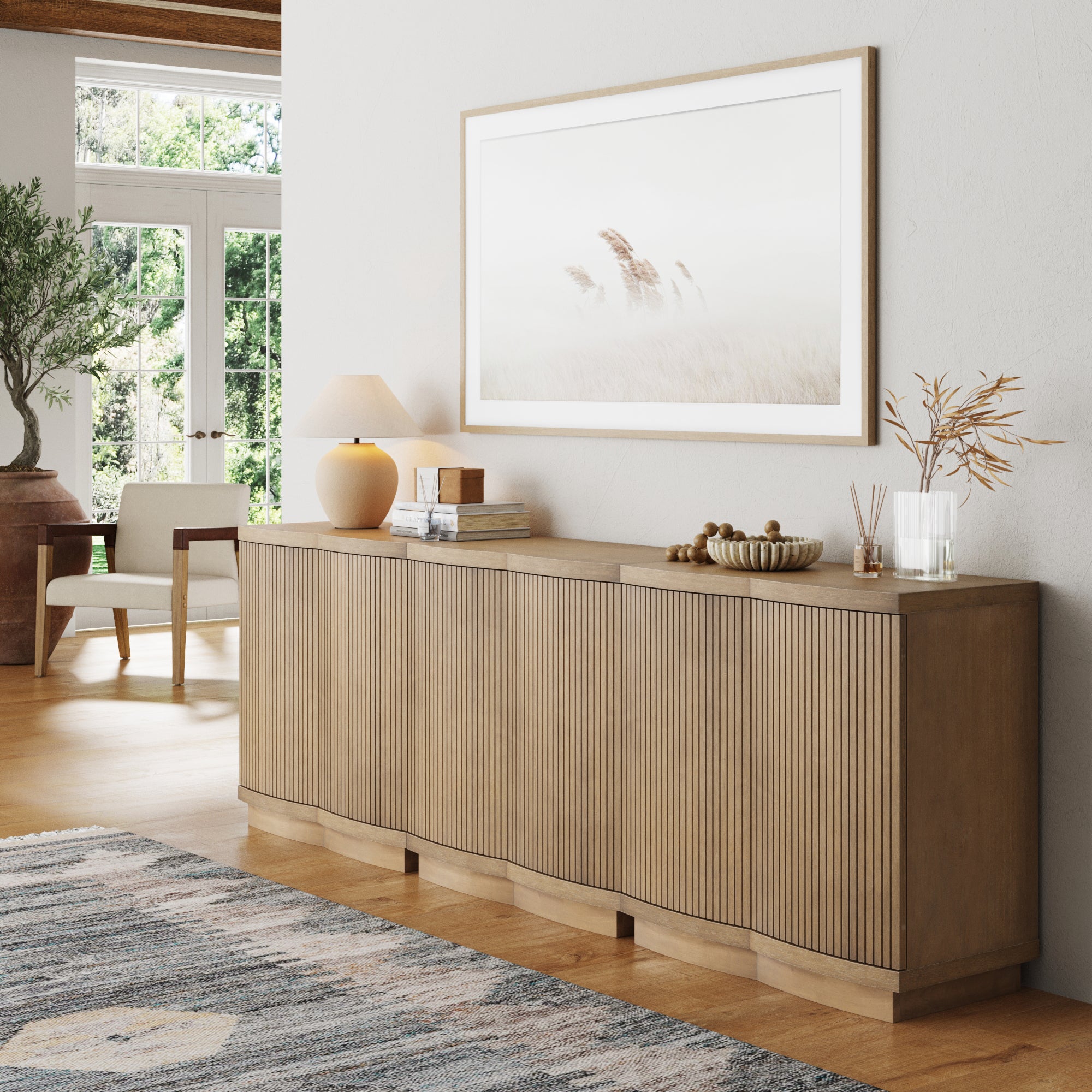 A modern living room showcases the Nathan James Mid-Century Modern Fluted Console or TV Stand Light Brown (Set of 3), with a table lamp and decor, a large framed nature photo above, and sunlight streaming onto a patterned rug on the wooden floor.