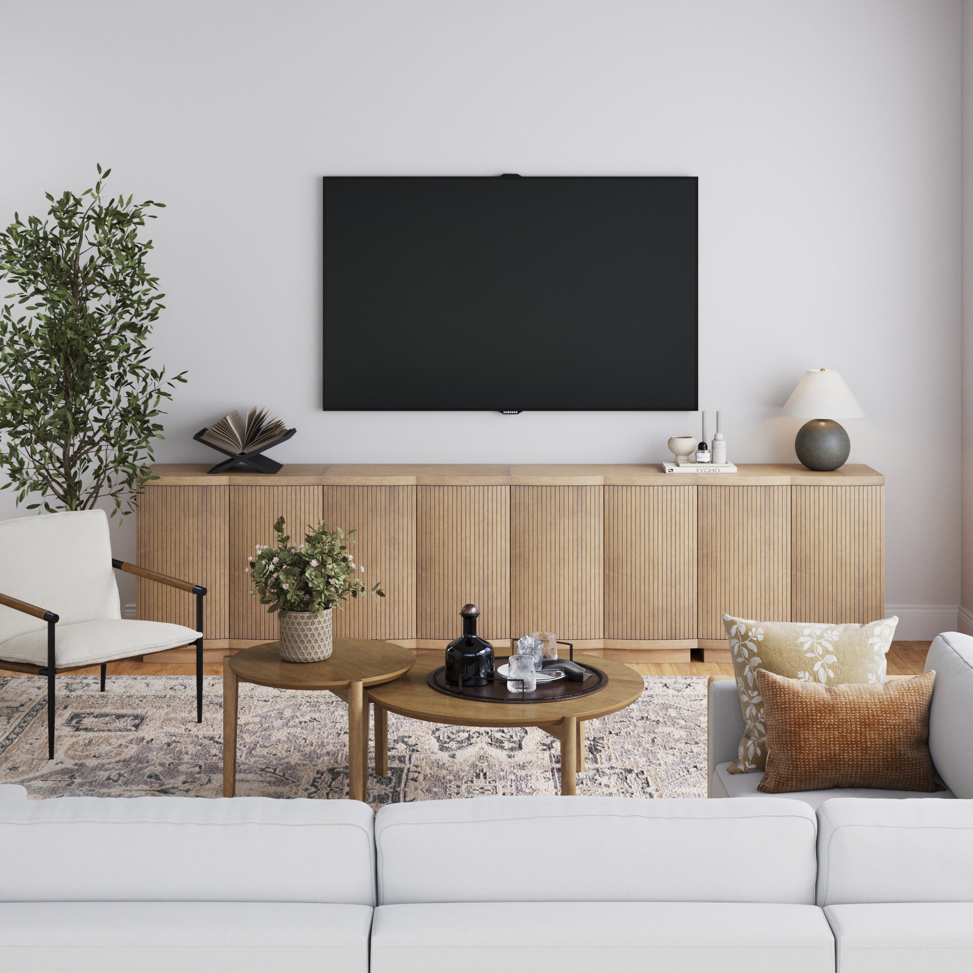 A modern living room features a white sofa with patterned pillows, two round wood coffee tables, and a large TV mounted above the Nathan James Mid-Century Modern Fluted Credenza TV Stand (Set of 4). A potted plant and beige armchair sit on the left.