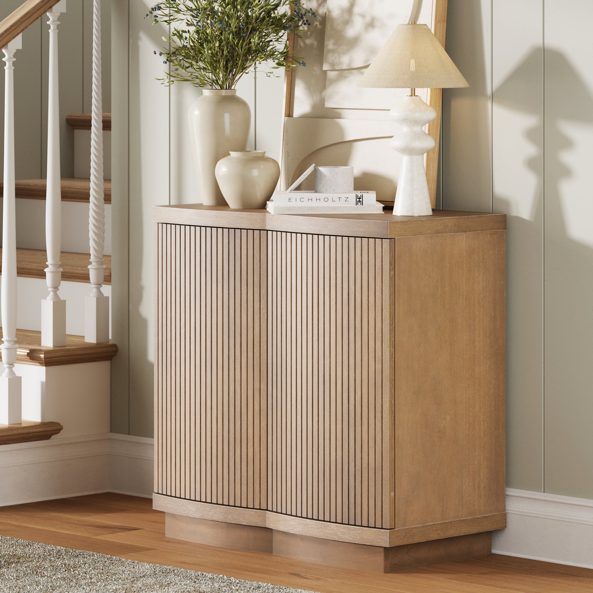 The Nathan James Mid-Century Modern Fluted Accent Cabinet in light brown stands by paneled walls and wooden stairs, topped with a potted plant, two white vases, a book, and a modern white lamp.