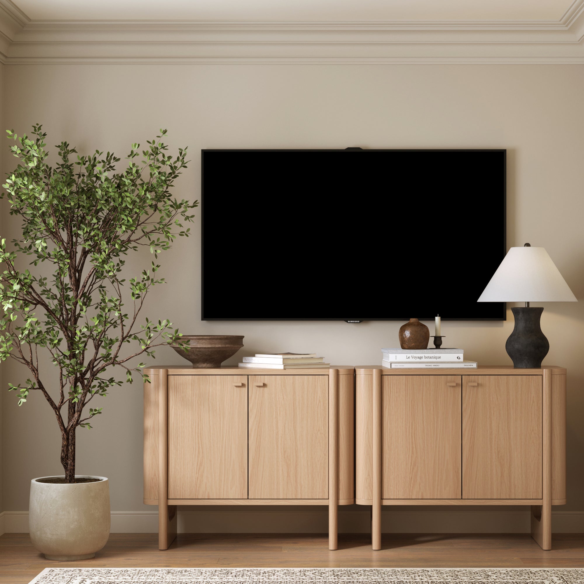 A modern living room features a large potted plant, books, decor, a table lamp, and wall-mounted TV, with two Nathan James Mid-Century Modern Solid Wood Accent Cabinets or TV Stands in Raw Oak set against a light beige wall.