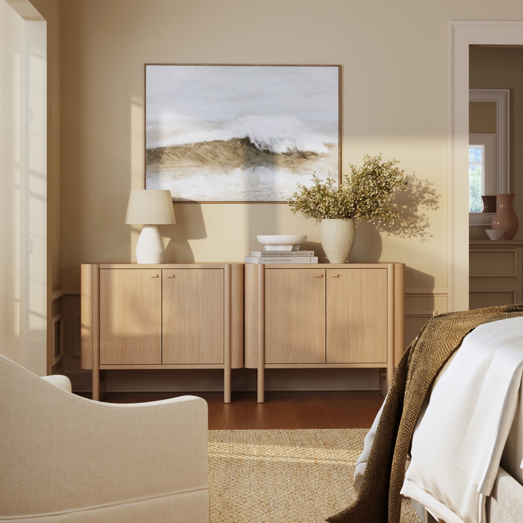 A cozy living room corner features two Nathan James Mid-Century Modern Solid Wood Accent Cabinets in Raw Oak, a lamp, stacked books, and a large flower vase. An ocean wave painting hangs above, filling the neutral space with soft light.