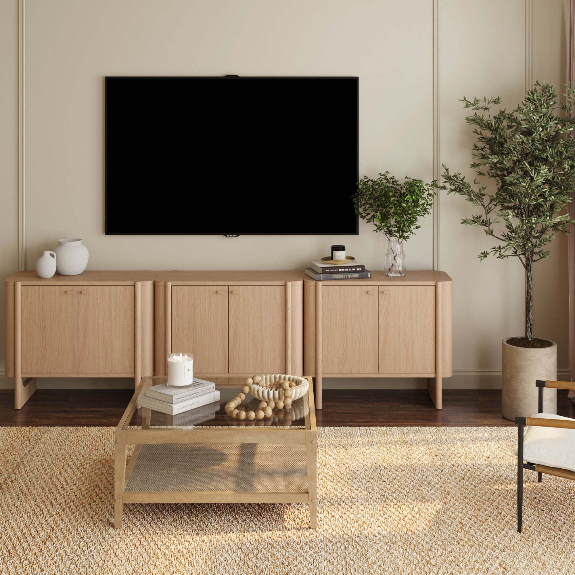 A modern living room features a wall-mounted TV, Nathan James Mid-Century Modern Solid Wood Accent Cabinet or TV Stand Raw Oak (Set of 3), decorative vases, books, green plants, a beige rug, and glass coffee table in cozy neutral tones.