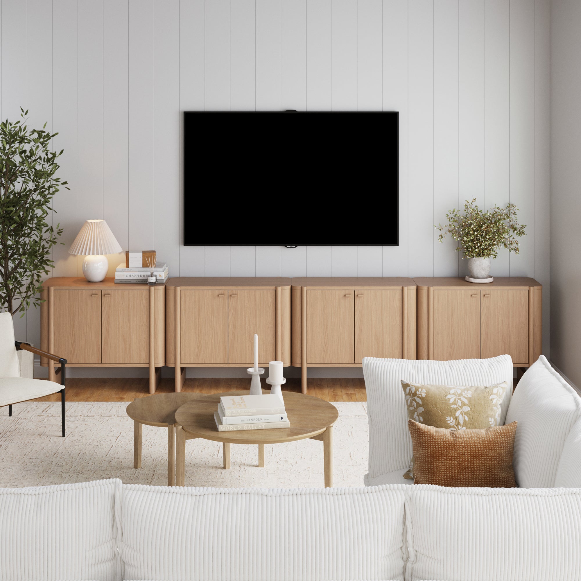 Modern living room with a white sofa, wooden coffee table, Nathan James Mid-Century Modern Solid Wood Credenza TV Stand in raw oak (set of 4) along the wall, large wall-mounted TV, decorative vases, plants, and soft neutral tones.