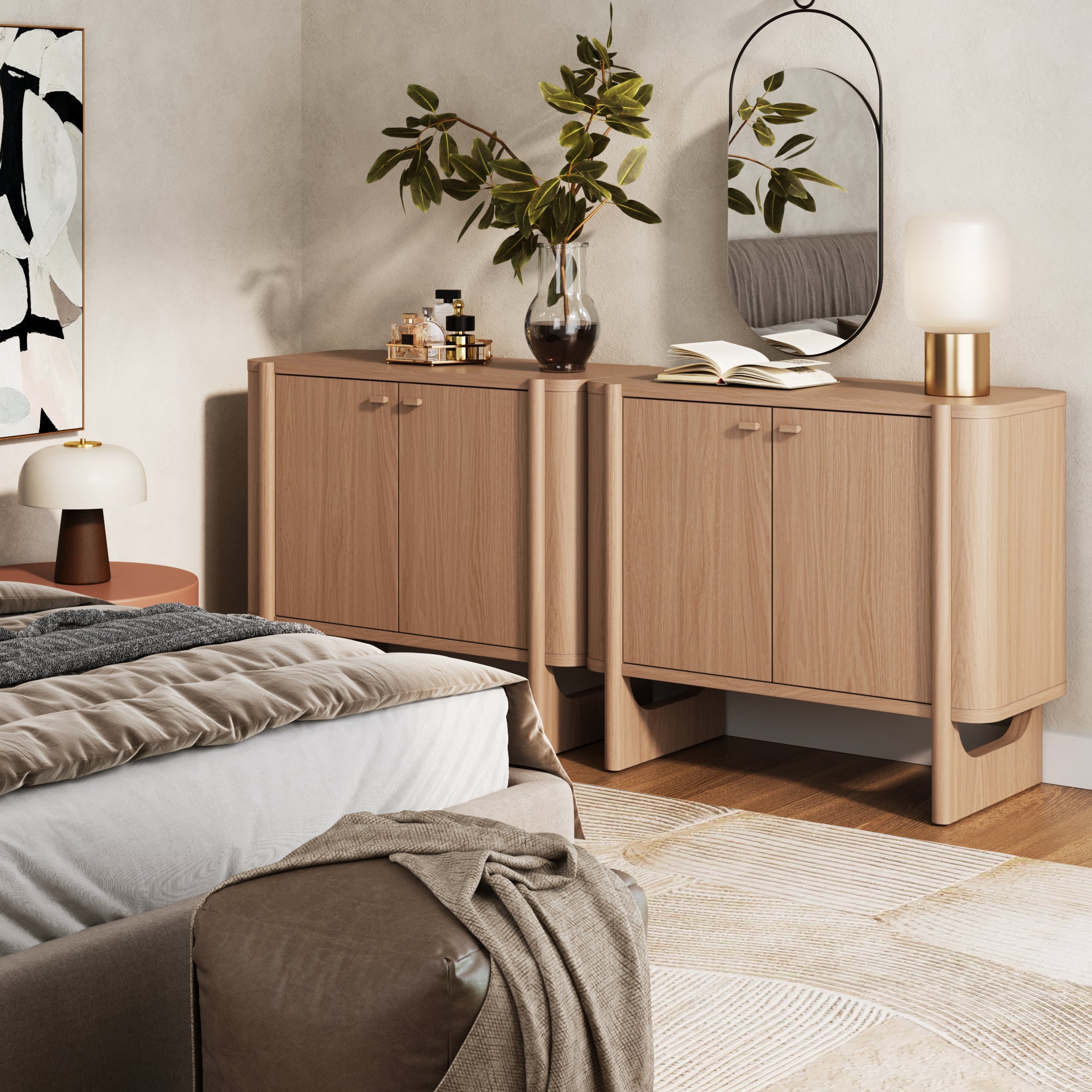 A modern bedroom features a wooden sideboard with plants, a lamp, books, and decor in front of a round mirror. Part of a bed, a brown ottoman, and soft neutral-toned furnishings are also visible.