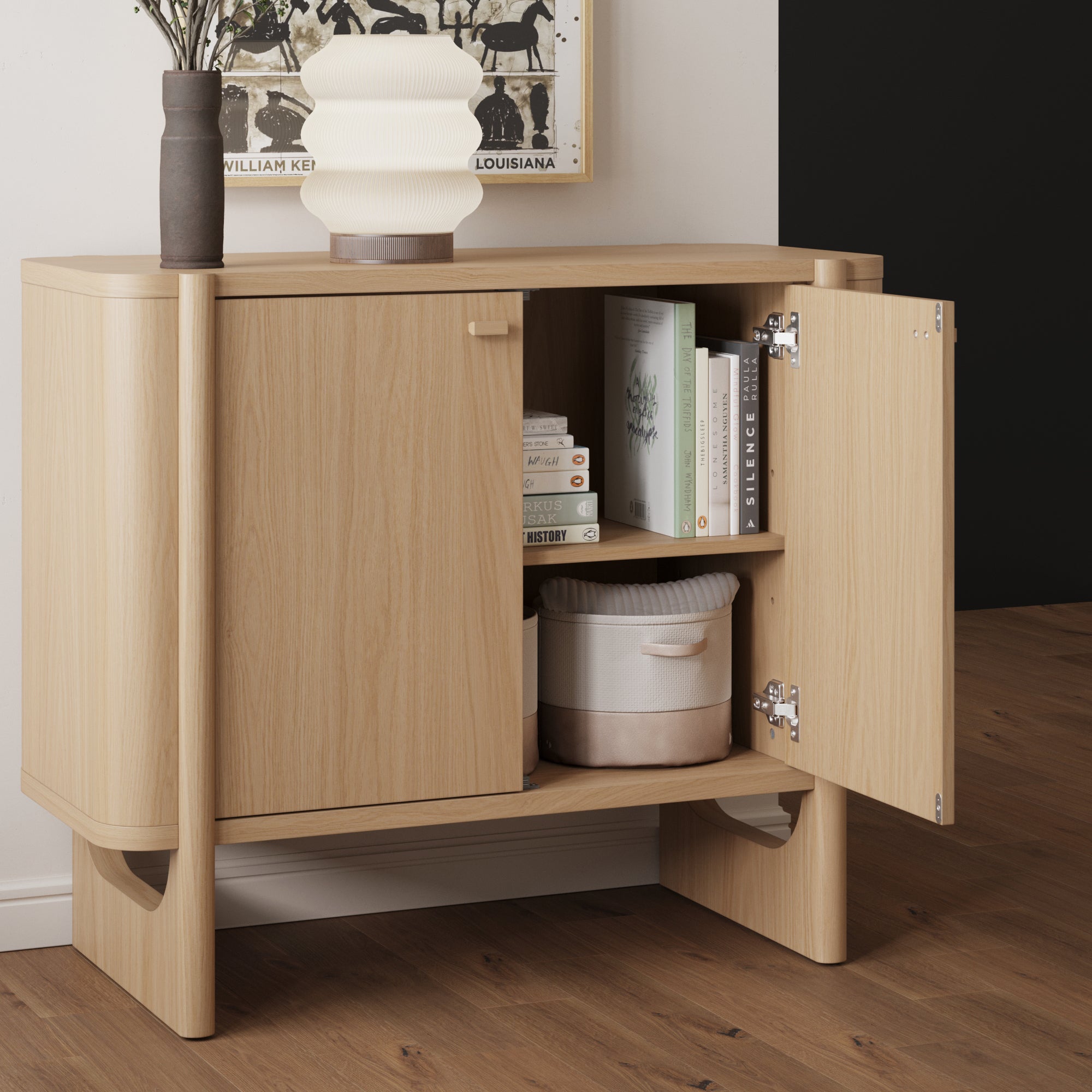 A light wood cabinet stands on a wooden floor, with one door open revealing books and a round storage basket inside. On top, there are two vases—one with flowers and one sculpted—and a framed artwork on the wall behind.