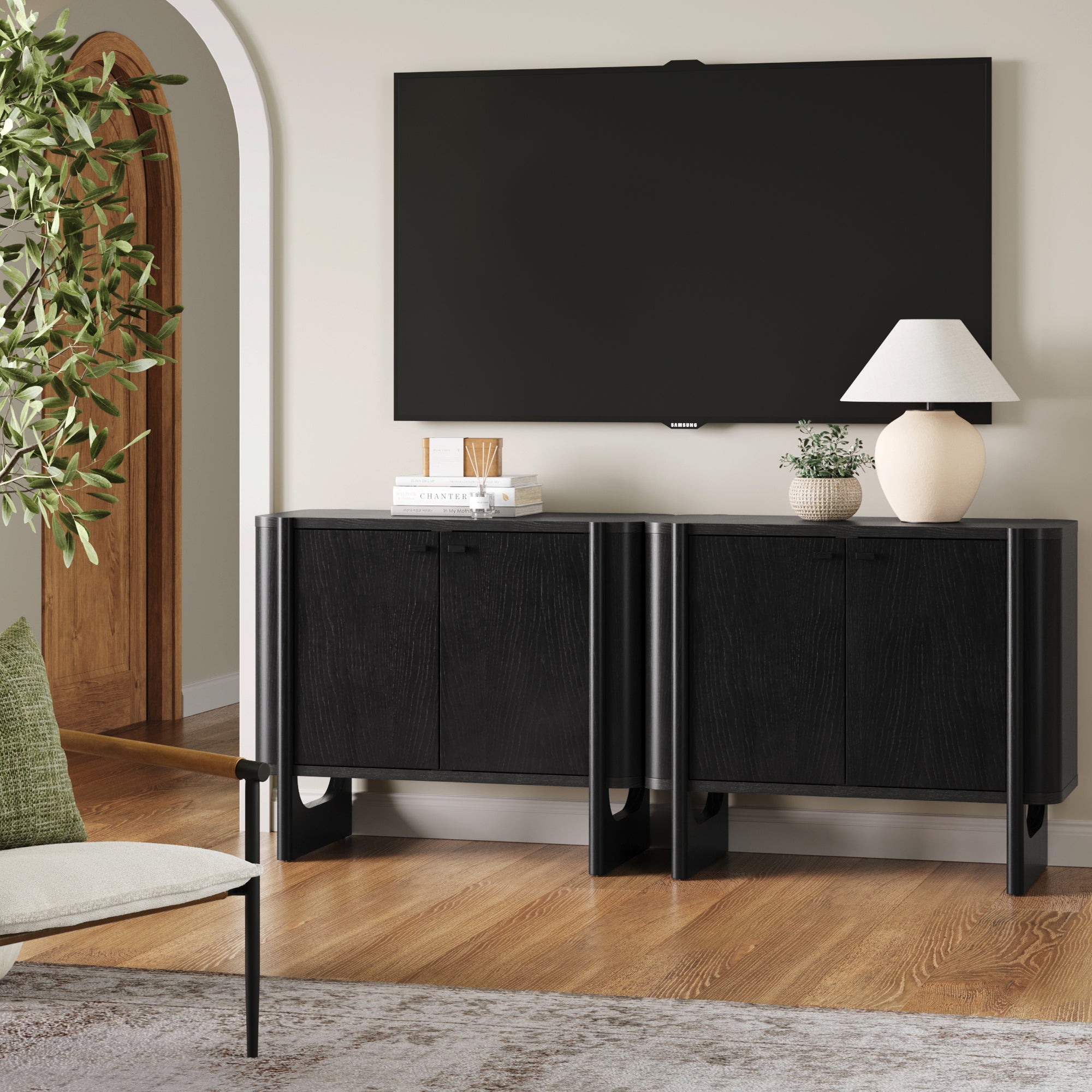 A modern living room features two black Nathan James Mid-Century Modern Solid Wood Accent Cabinets, a wall-mounted flat-screen TV, white table lamp, decorative books, beige vase, green plant, and a white chair on a wooden floor.