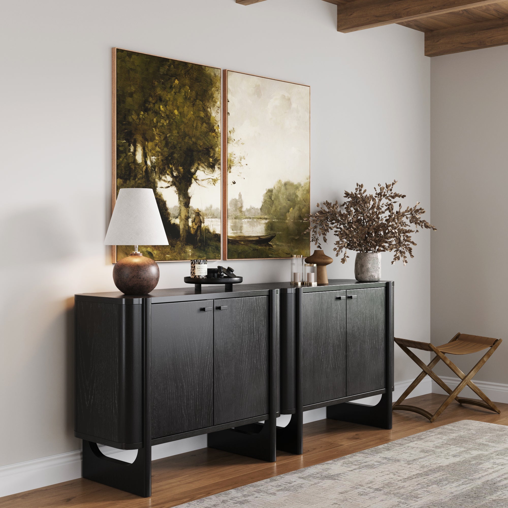 A pair of Nathan James Mid-Century Modern Solid Wood Accent Cabinets in black display a lamp, vases, and dried branches. Two landscape paintings hang above, with a folding wooden stool nearby on the hardwood floor.