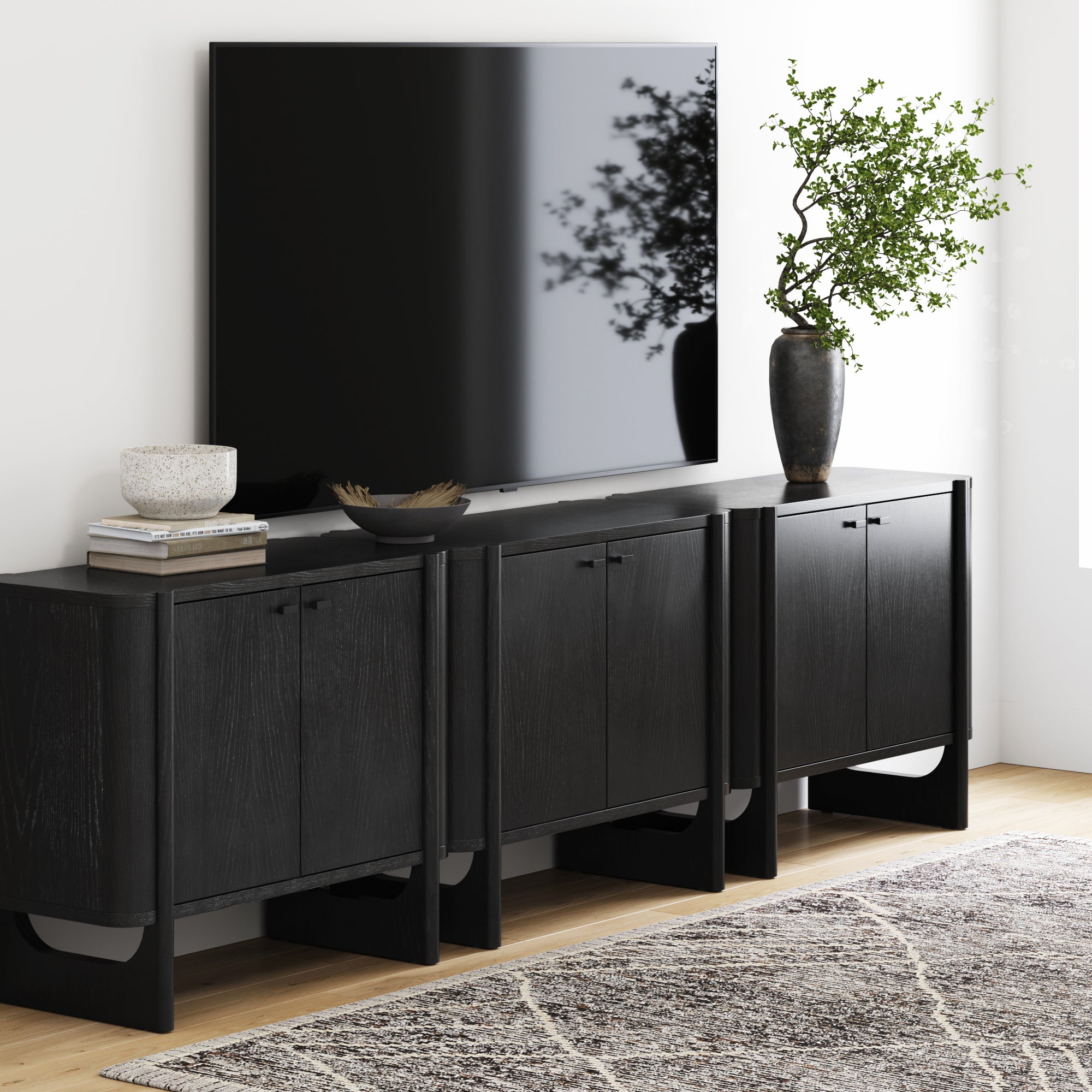 A modern living room features a large TV above the Nathan James Mid-Century Modern Solid Wood Accent Cabinet or TV Stand in black (set of 3), decorated with a vase, bowls, and books. A gray patterned rug covers the floor.