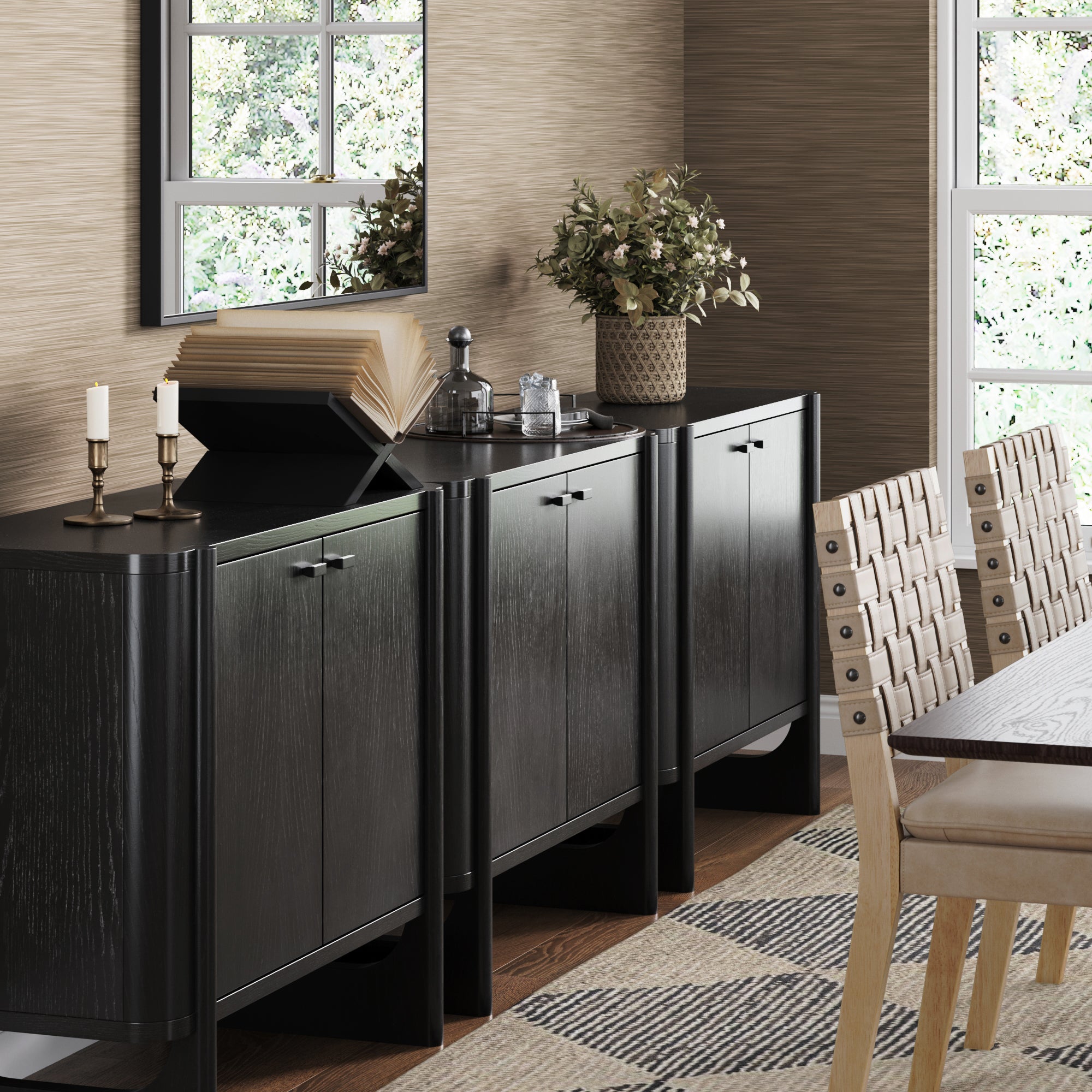 A modern dining room showcases the Nathan James Mid-Century Modern Solid Wood Accent Cabinet or TV Stand (Set of 3, Black), holding candles, a book, decanters, and a plant. Light wood chairs and a patterned rug sit beneath a window and wall mirror.