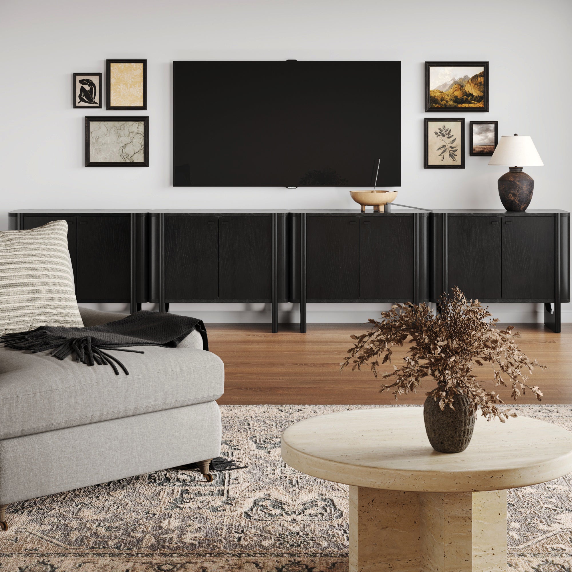 A modern living room featuring a gray sofa, round beige coffee table with dried flowers, large wall-mounted TV, four Nathan James Mid-Century Modern Solid Wood Credenza TV Consoles in black, framed art, lamp, wood floor, and patterned rug.