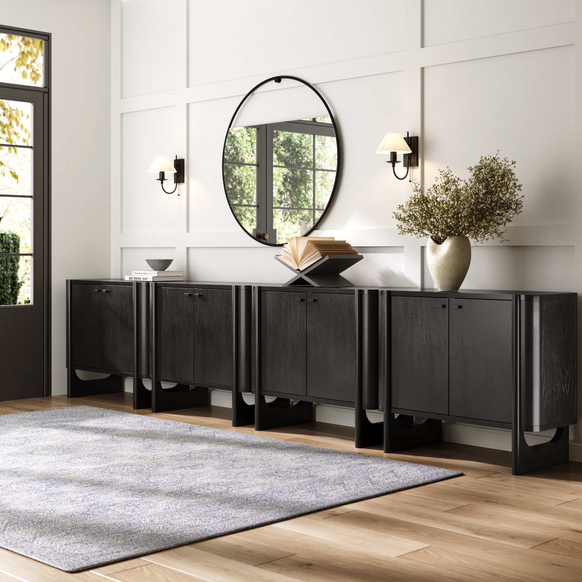 A modern entryway showcases the Nathan James Mid-Century Modern Solid Wood Credenza TV Console in black (set of 4), a large round mirror, two wall sconces, greenery in a vase, decorative books, and a bowl—all against white paneled walls and wood flooring.