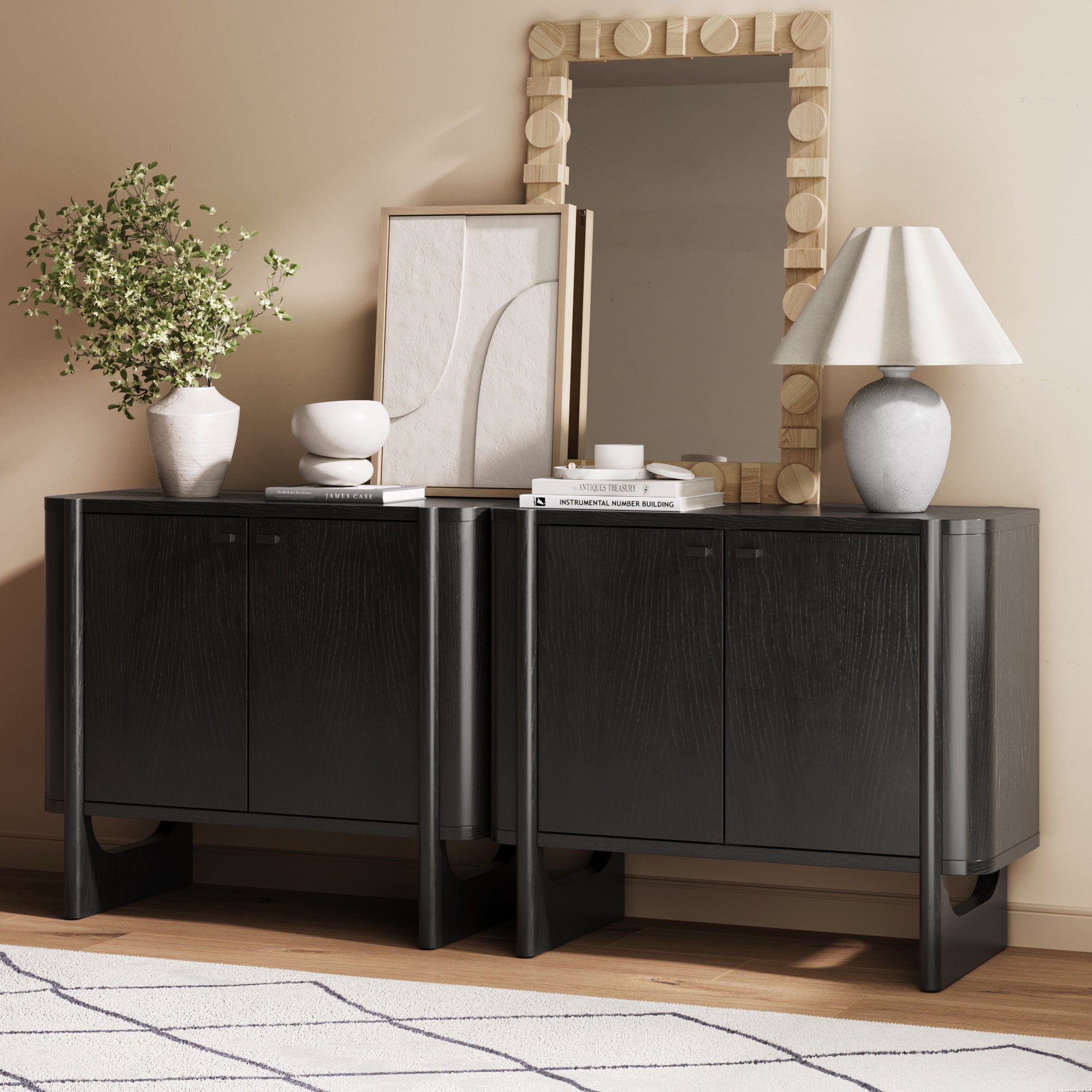 A modern room with two black cabinets, a large wooden-framed mirror, decorative vases with greenery, abstract art, stacked books, and a lamp on top; a light rug with a geometric pattern covers the floor.