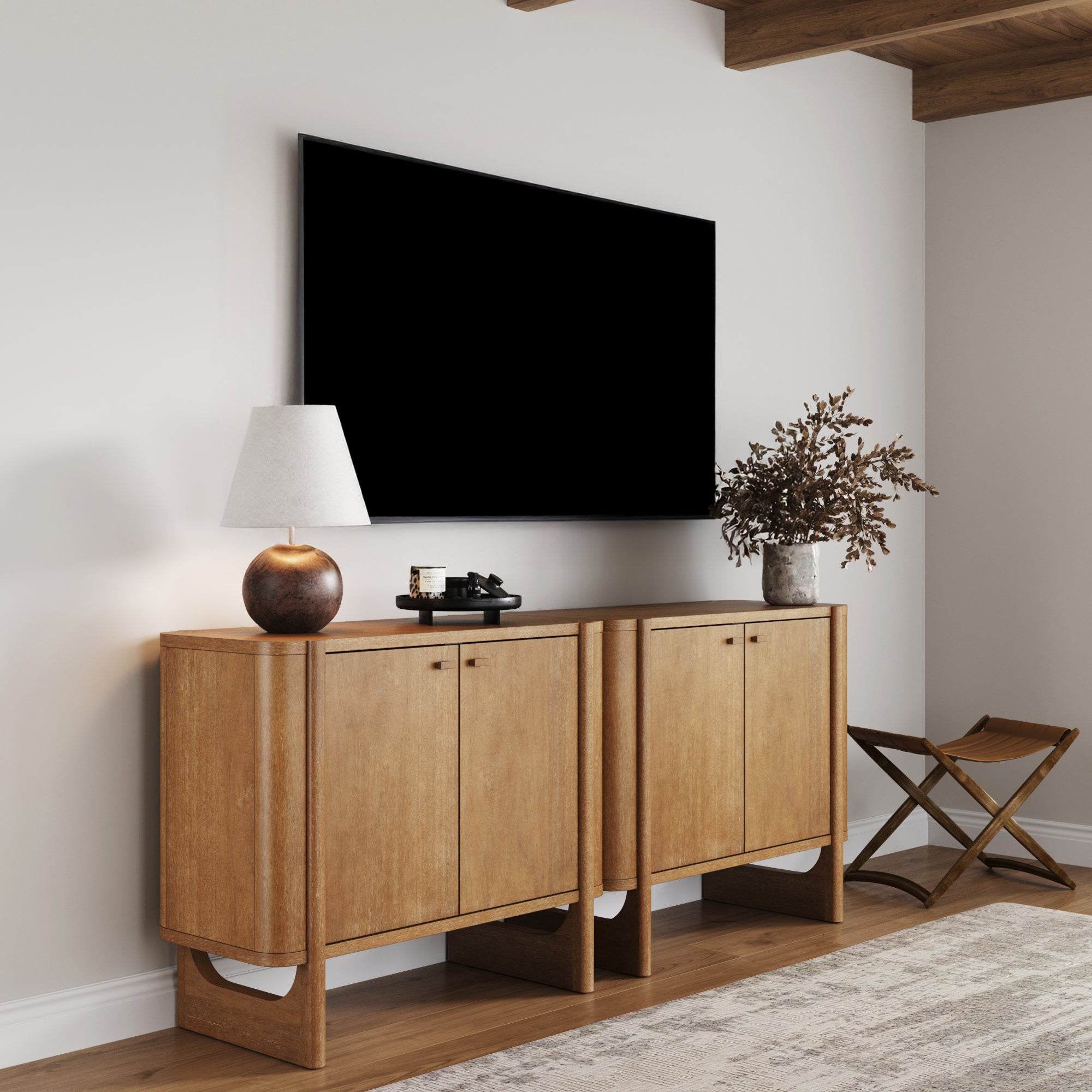 A modern living room with a large wall-mounted TV above two Nathan James Mid-Century Modern Solid Wood Accent Cabinets or TV Stands in light brown, plus a lamp, decor, vase with dried branches, folding stool, and rug on hardwood floors.