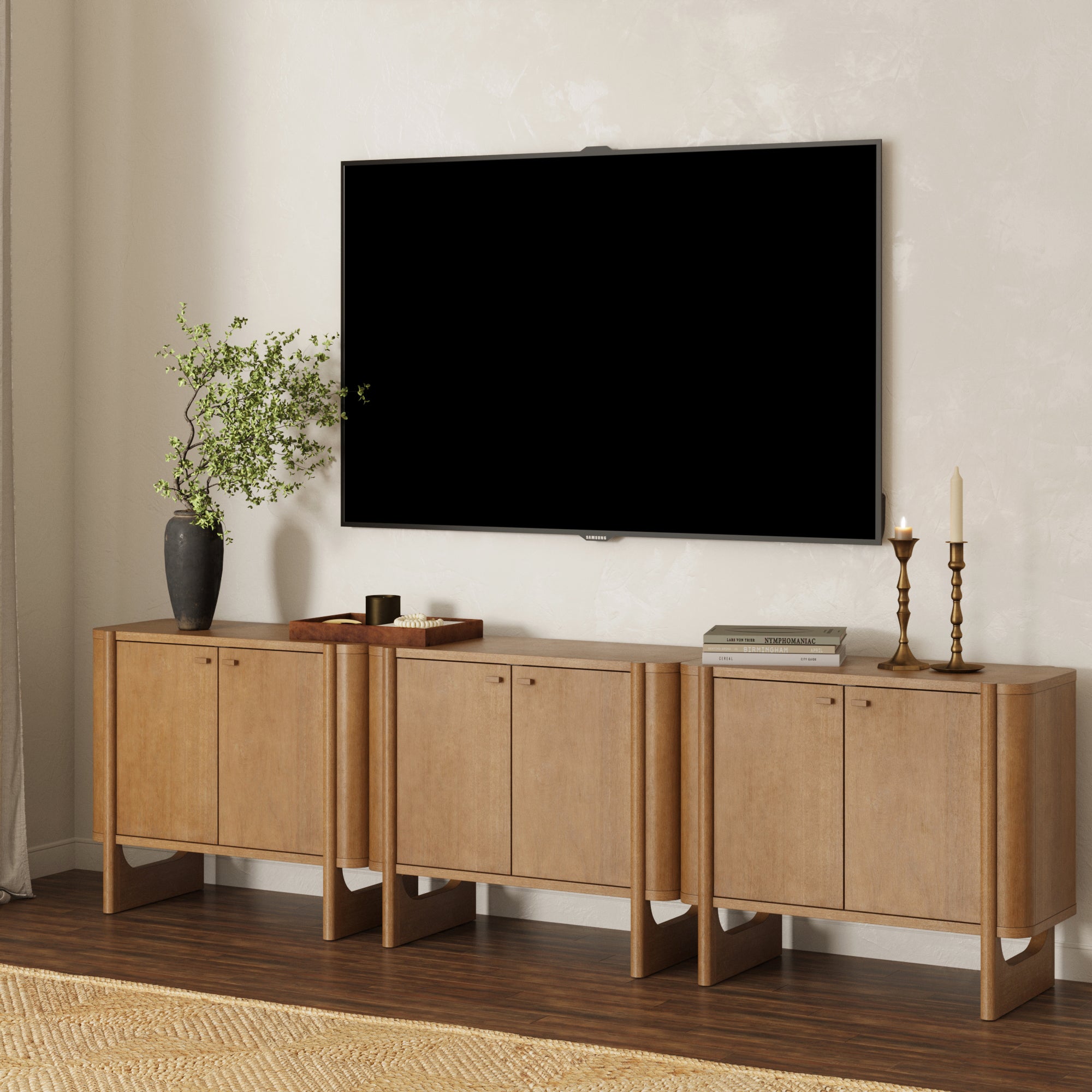 A modern living room features a large flat-screen TV above the Nathan James Mid-Century Modern Solid Wood Accent Cabinet or TV Stand (Set of 3), light brown, styled with a vase, tray, books, and candlesticks.