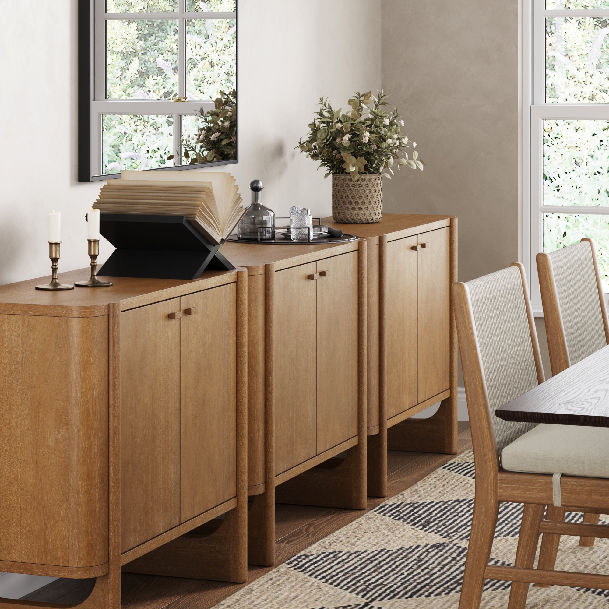The Nathan James Mid-Century Modern Solid Wood Accent Cabinet or TV Stand (Set of 3, Light Brown) holds books, a plant, glassware, and candlesticks in a dining room with a window, mirror, and beige chairs around the table.