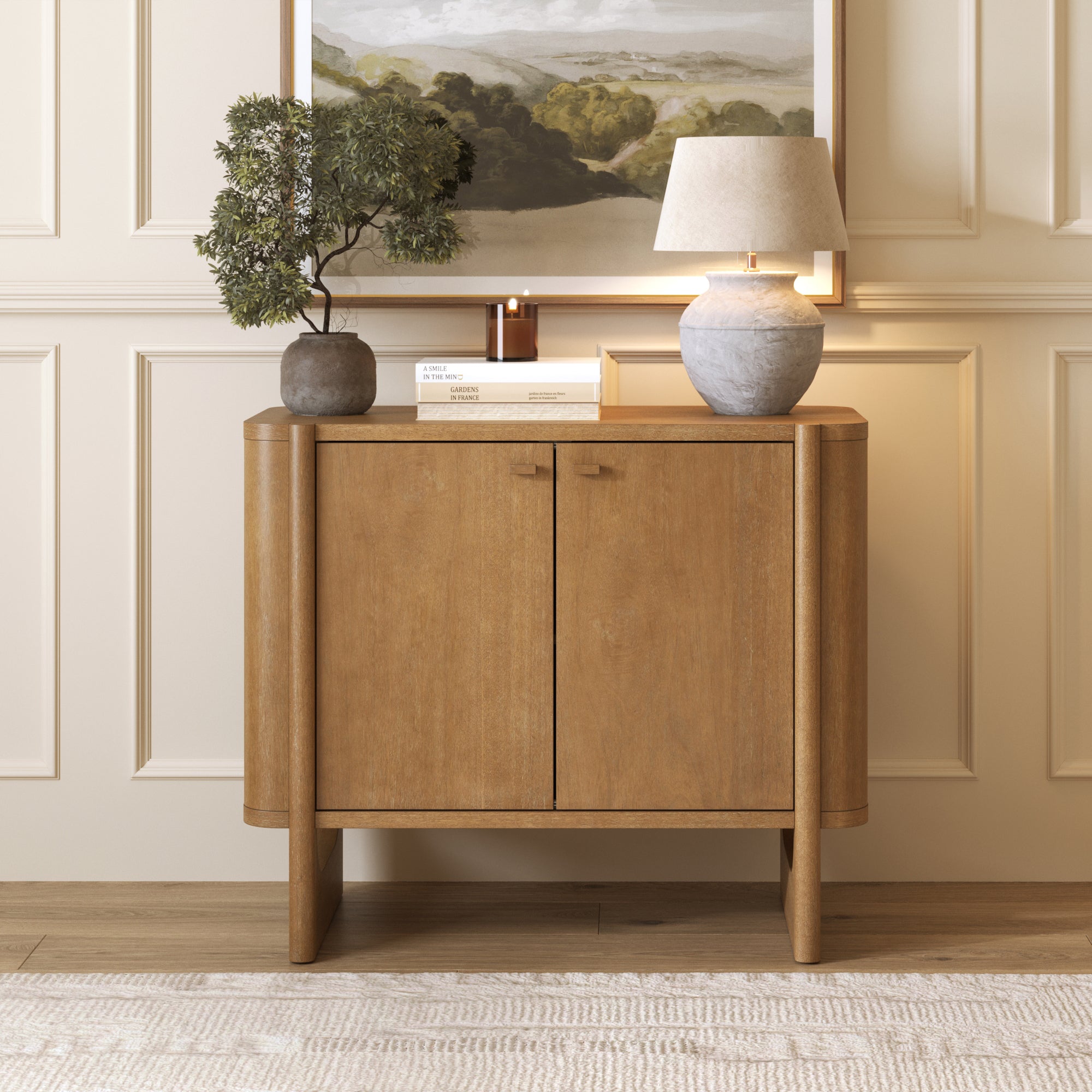 A Nathan James Mid-Century Modern Solid Wood Accent Cabinet in light brown with two doors stands against a cream paneled wall. On top are a potted plant, beige-shaded lamp, and candle; a landscape painting hangs above and a neutral rug lies underfoot.