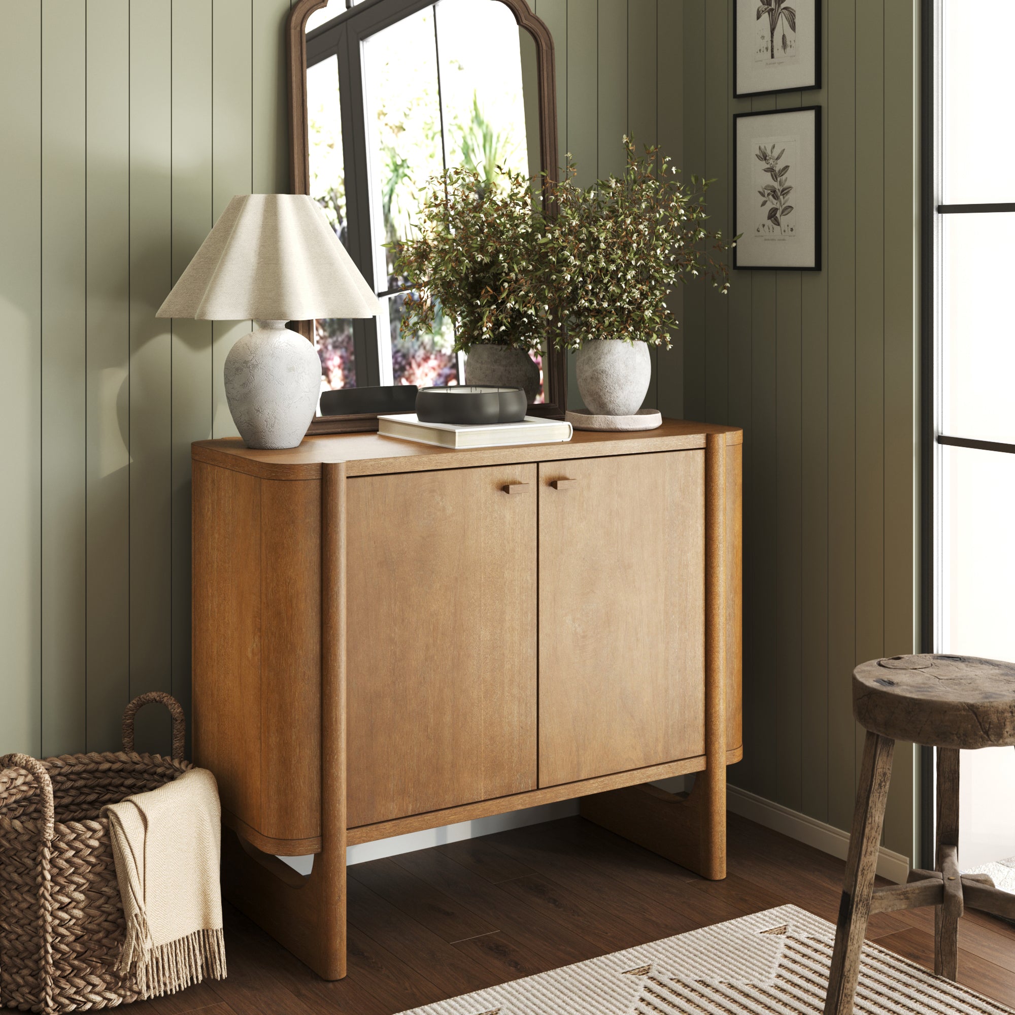 A Nathan James Mid-Century Modern Solid Wood Accent Cabinet in light brown with two doors stands against a green paneled wall, topped with a white lamp, vases of greenery, a tray, and mirror; nearby are a wicker basket and wooden stool on the floor.