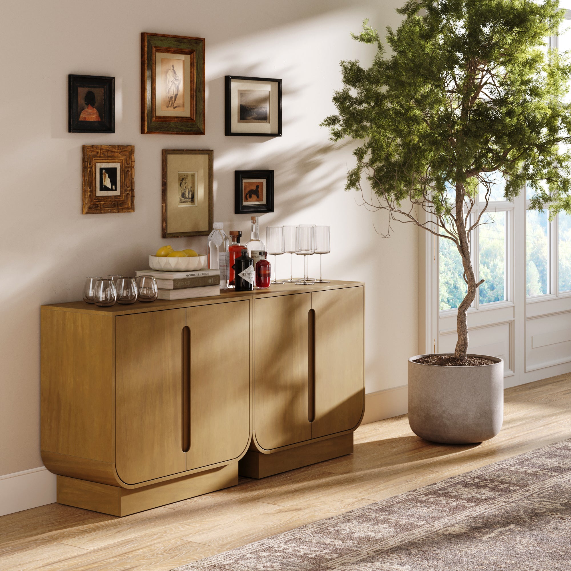A modern living space featuring two Nathan James Veneer Arched Accent Cabinets in light brown, used as storage or TV stands, topped with glassware, bottles, and a bowl of lemons, alongside a potted tree, framed wall art, and sunlit tall windows.