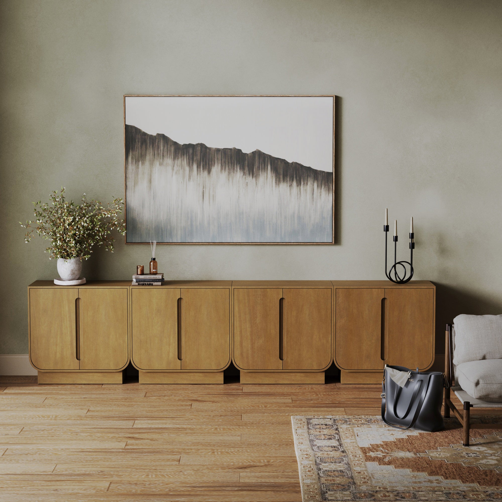 A modern living room featuring the Nathan James Veneer Arched Large Credenza Cabinet (Set of 4) in light brown, a vase with green foliage, books, two black candle holders, an abstract mountain painting, a patterned rug, and a gray handbag.