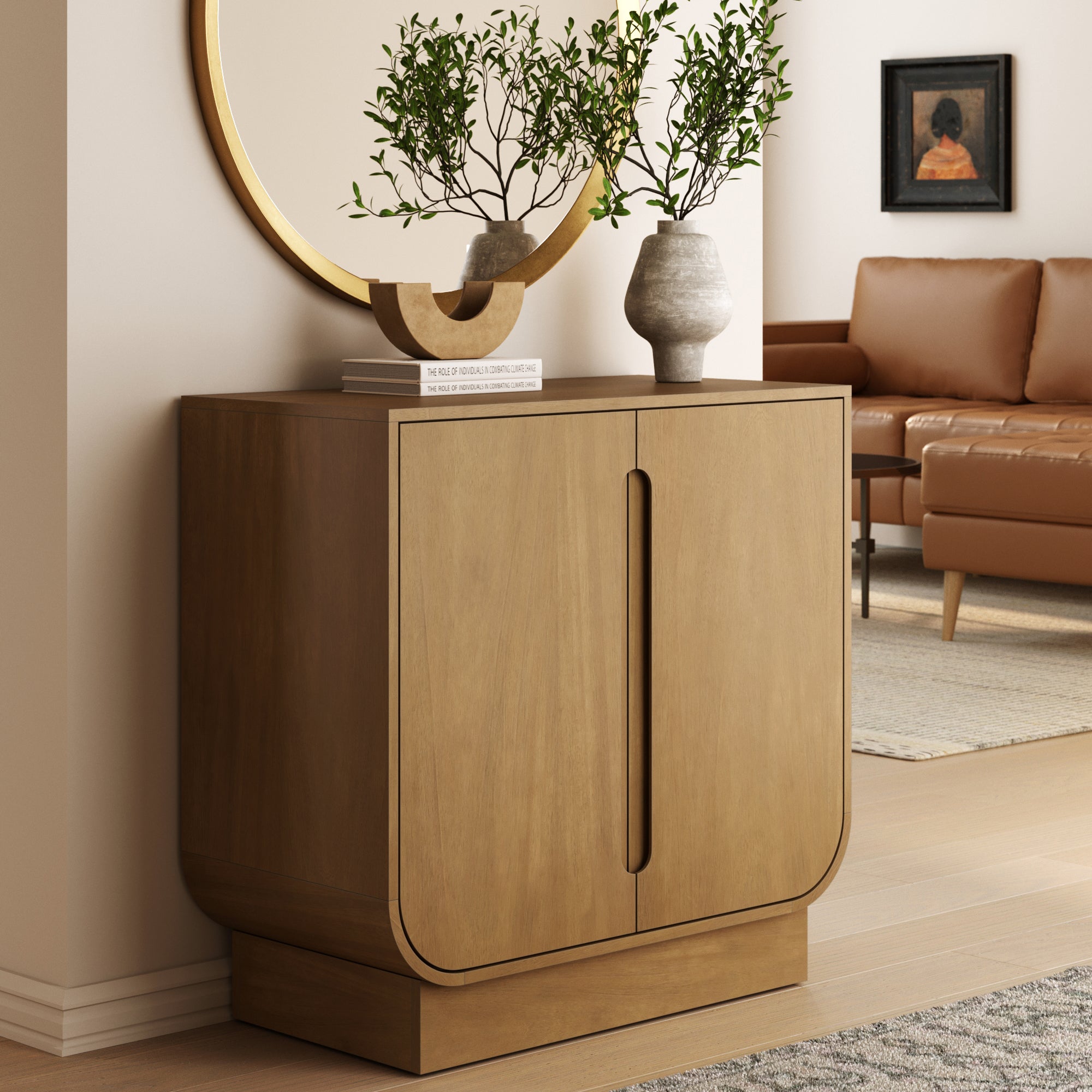 The Nathan James Wood Veneer Arched Accent Cabinet in light brown features a modern mid-century design, styled with potted plants, a mirror, and a book. A brown leather sofa and framed portrait complete the room’s warm, minimalist look.