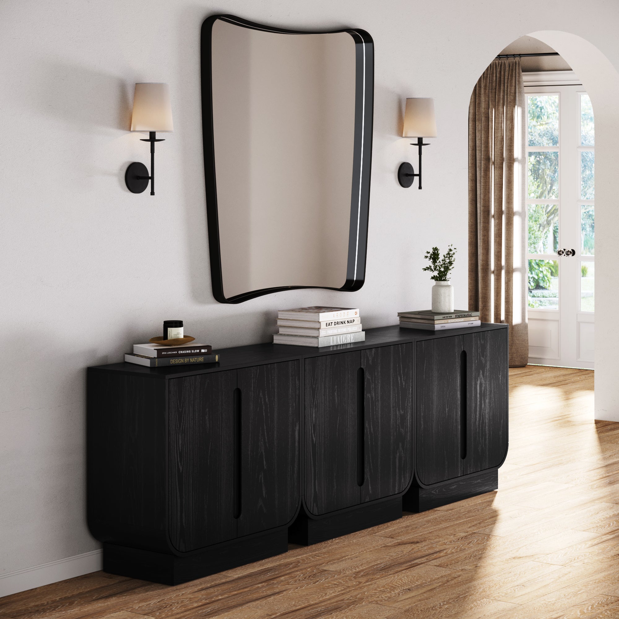A modern entryway showcases the Nathan James Veneer Arched Accent Cabinet or TV Stand in Black Oak (Set of 3), paired with a wavy mirror, wall sconces, books, a vase, a small plant, light wood flooring, and an arched doorway.