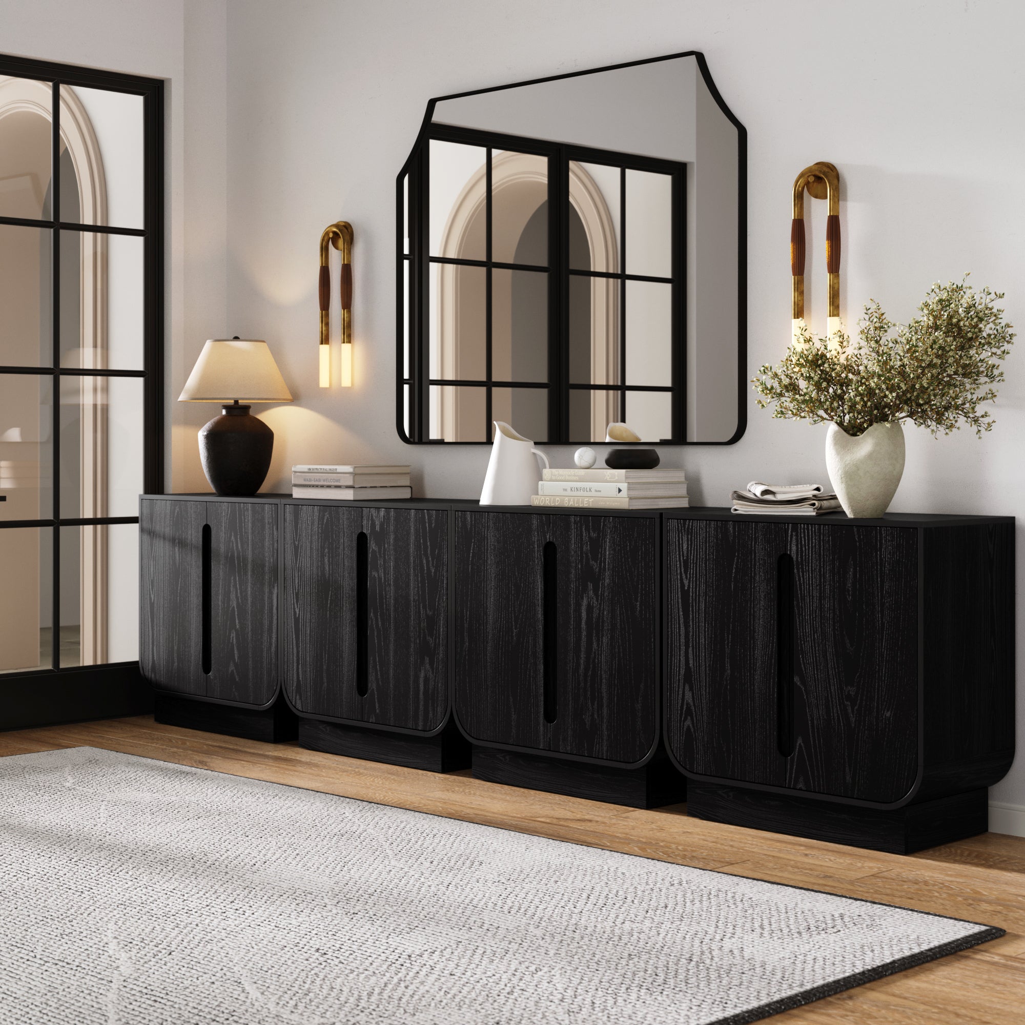 A modern entryway with the Nathan James Veneer Arched Large Credenza Cabinet in Black Oak (Set of 4), a large mirror, wall sconces, table lamp, books, decor accents, a greenery-filled vase, light walls, wood floors, and a textured gray rug.