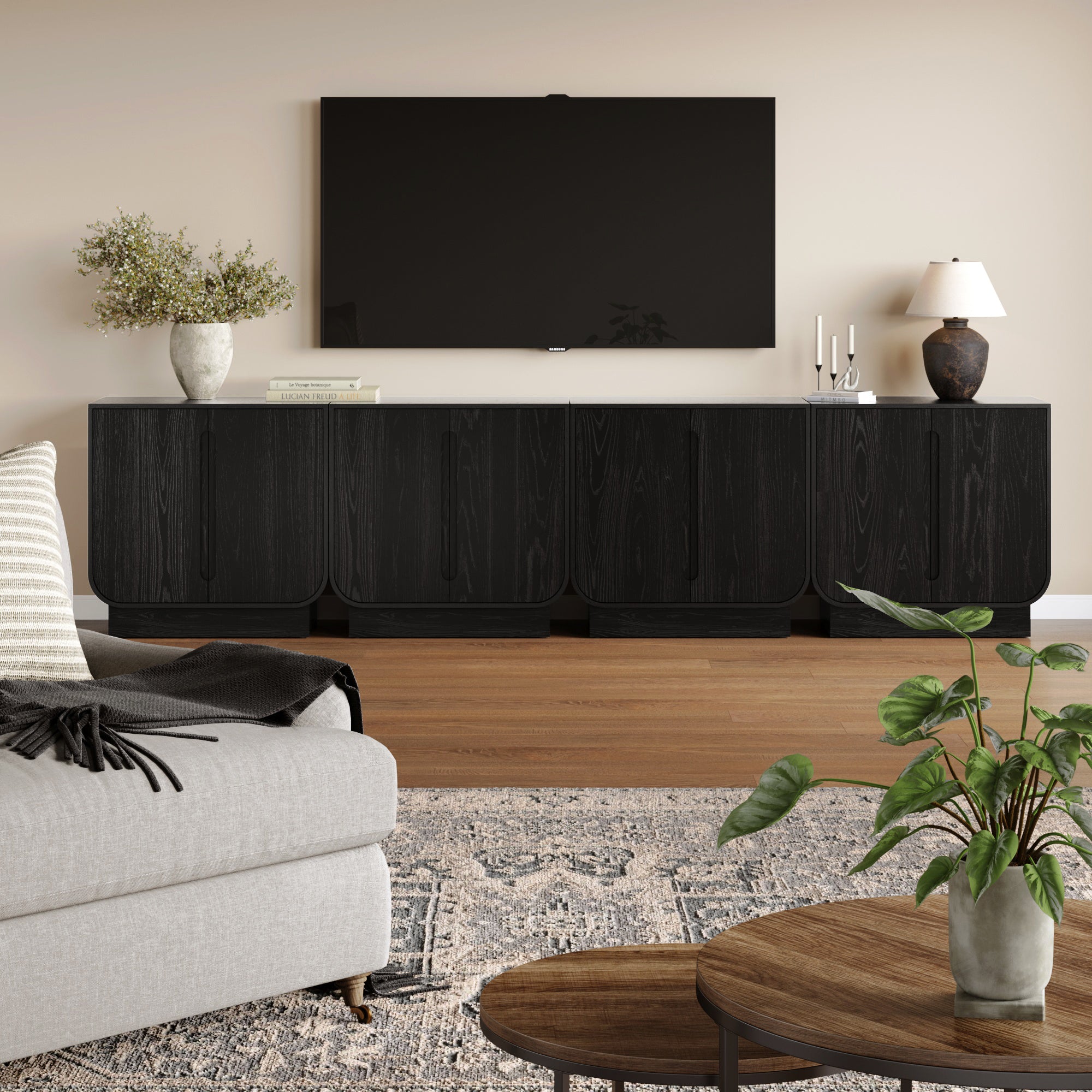 A modern living room showcases the Nathan James Veneer Arched Large Credenza Cabinet in Black Oak (Set of 4) beneath a wall-mounted TV, styled with greenery, books, and candles. A beige sofa faces a round table; a patterned rug completes the look.