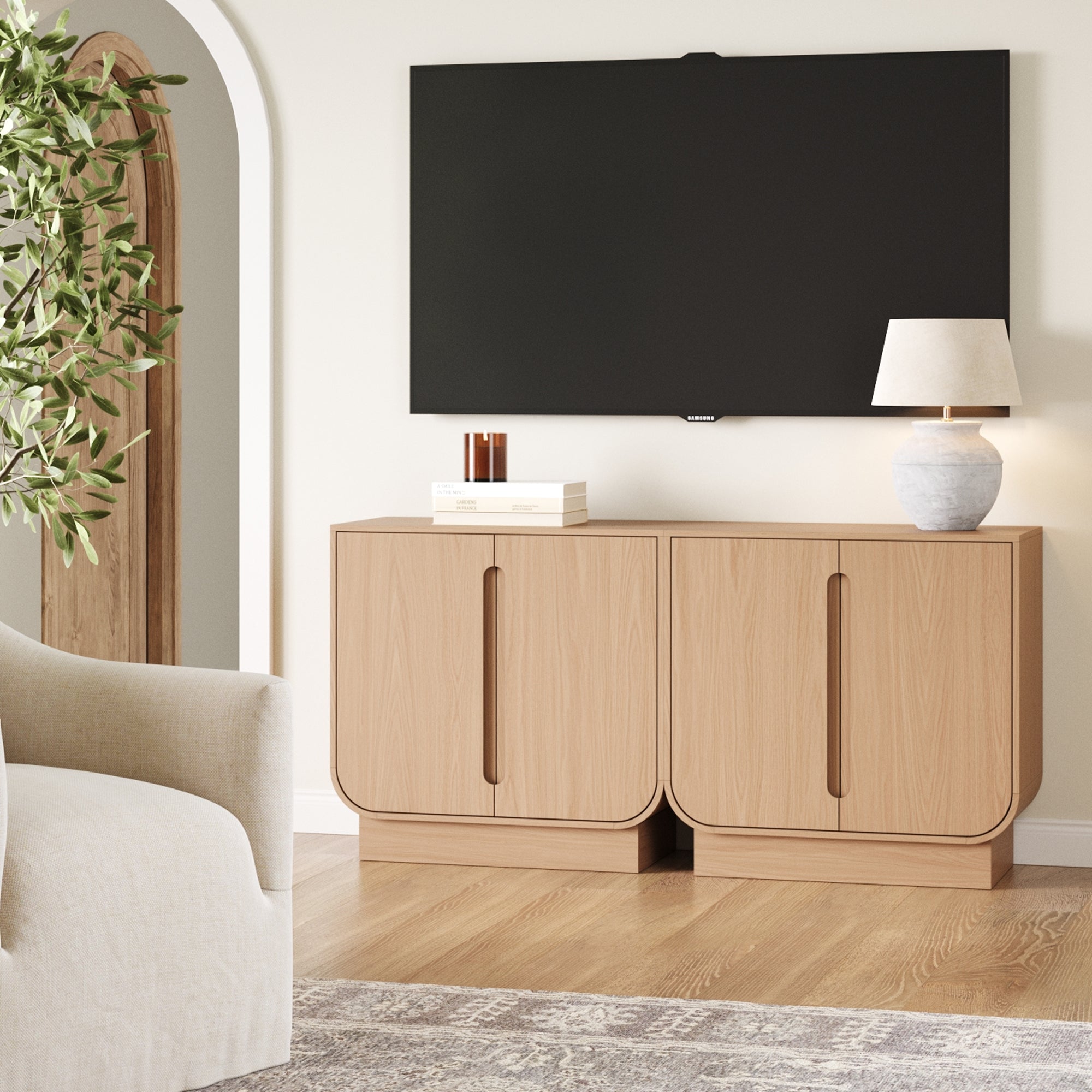 A modern living room showcases the Nathan James Veneer Arched Accent Cabinet or TV Stand in Raw Oak (Set of 2), paired with a lamp and books beneath a wall-mounted TV, plus a leafy plant by the archway and part of a light sofa and rug in view.